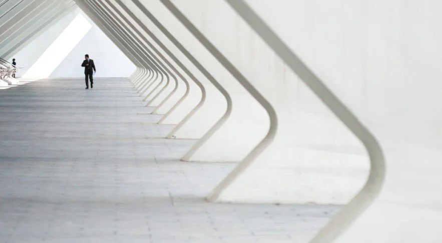 Un homme en costume marche dans un bâtiment moderne avec une architecture minimaliste, lignes épurées et accents blancs.
