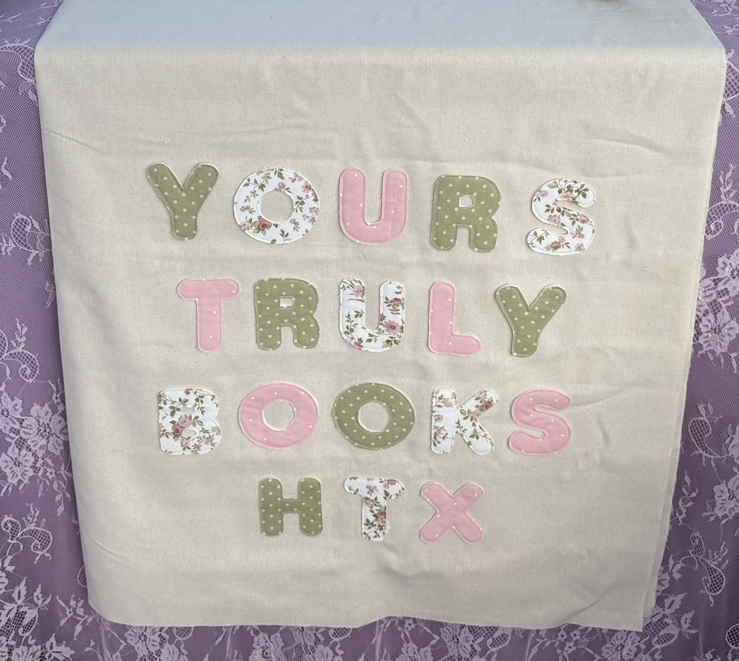 Fabric banner with flower-themed and polka dot letters spelling 'YOURS TRULY BOOKS HTX' on a white background, bordered by purple lace fabric.