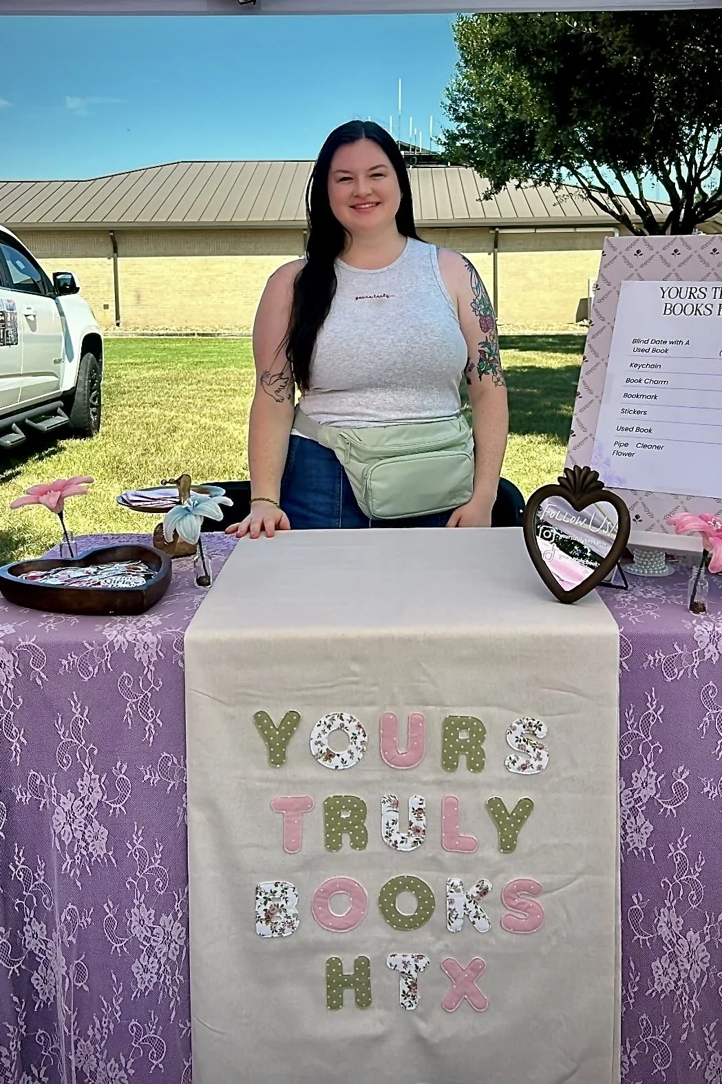 A woman standing behind a table at a book-themed event outdoors. The table has a sign saying 'Yours Truly Books' and decorative items. She has long dark hair, tattoos on her arms, and is wearing a sleeveless gray top with a light green fanny pack. Behind her, there are trees, a building, and a parked truck.
