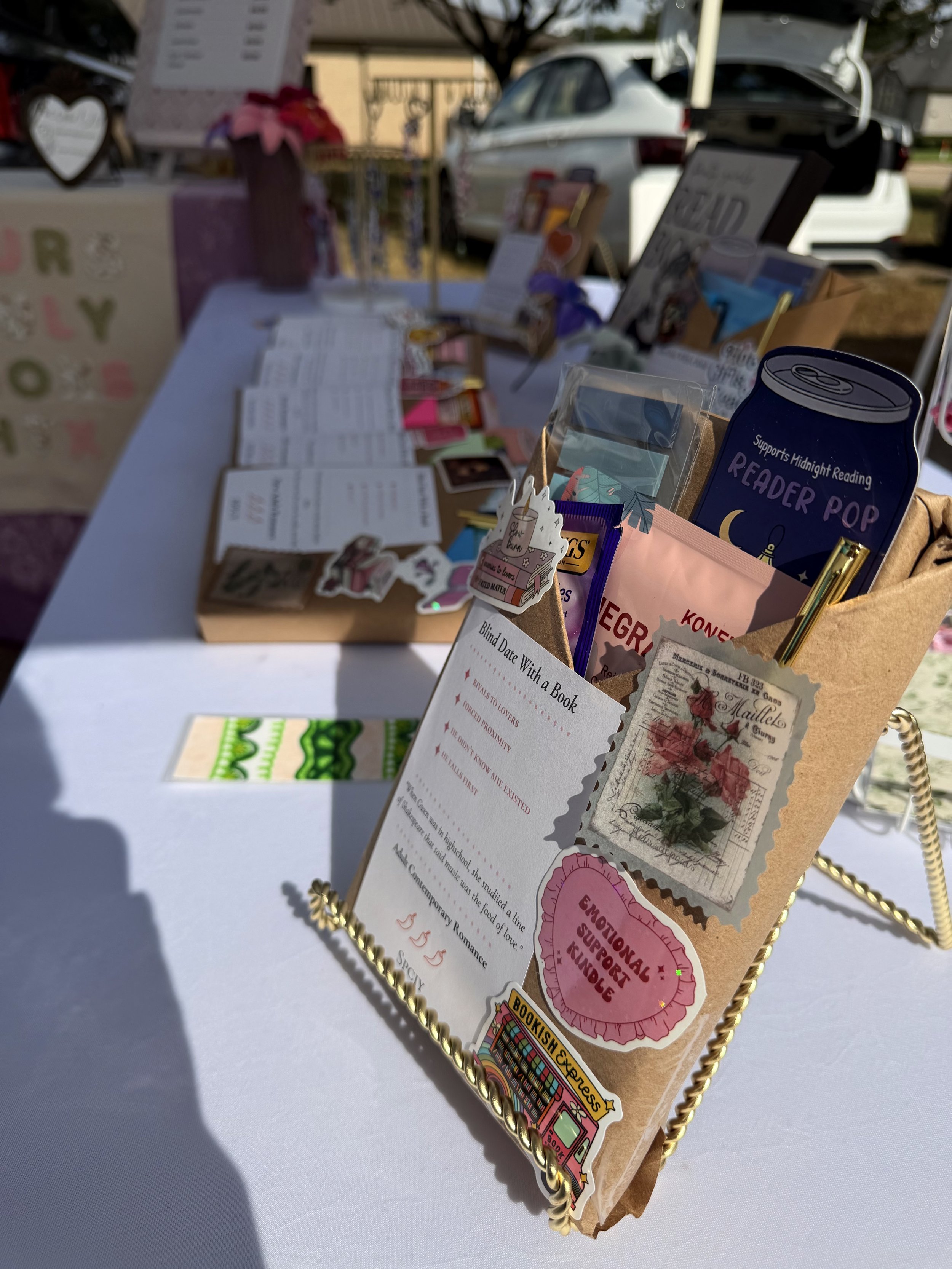 A display stand with various book-related items, flyers, and stickers, set up outdoors on a white table with a vehicle and trees in the background.