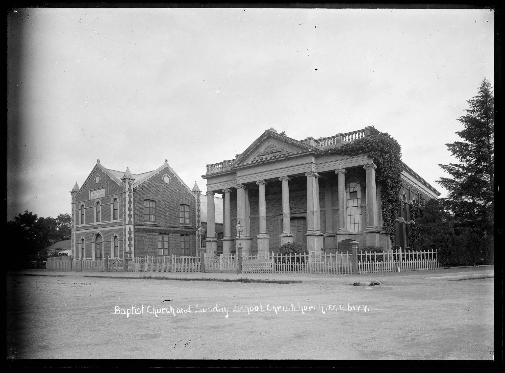 Black & white photograph of the Oxford Terrace Baptist Church (& sunday school). Brick building with classical styling: pediment, Corinthian columns, typanum. Wooden picket fence. Ivy growing on the building.