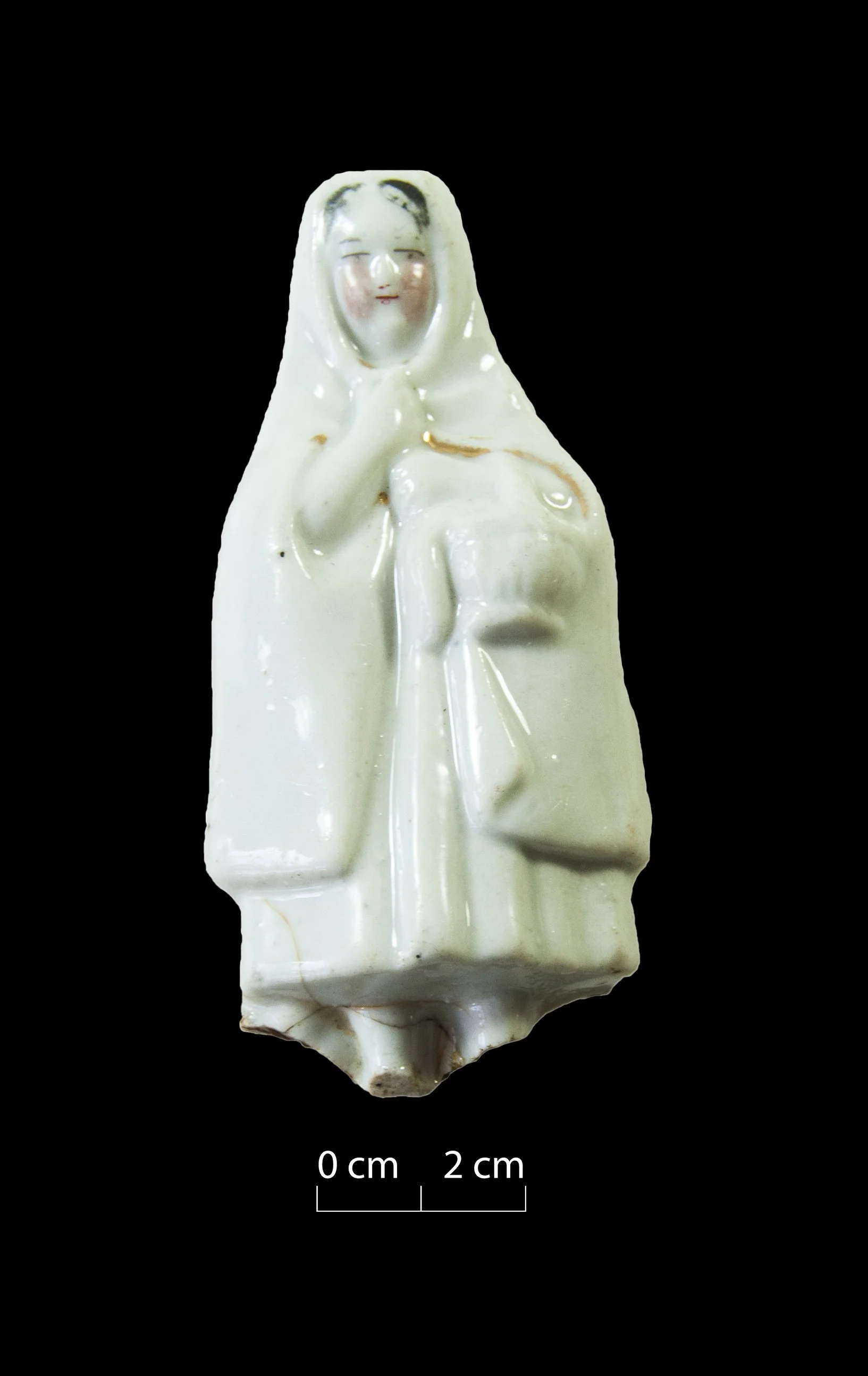 A porcelain figurine. Female. Largely white, but black hair, red cheeks, mouth and eyes painted on. Wears a cloak over her head, held in place by one hand. Carries a basket. Broken at the base.