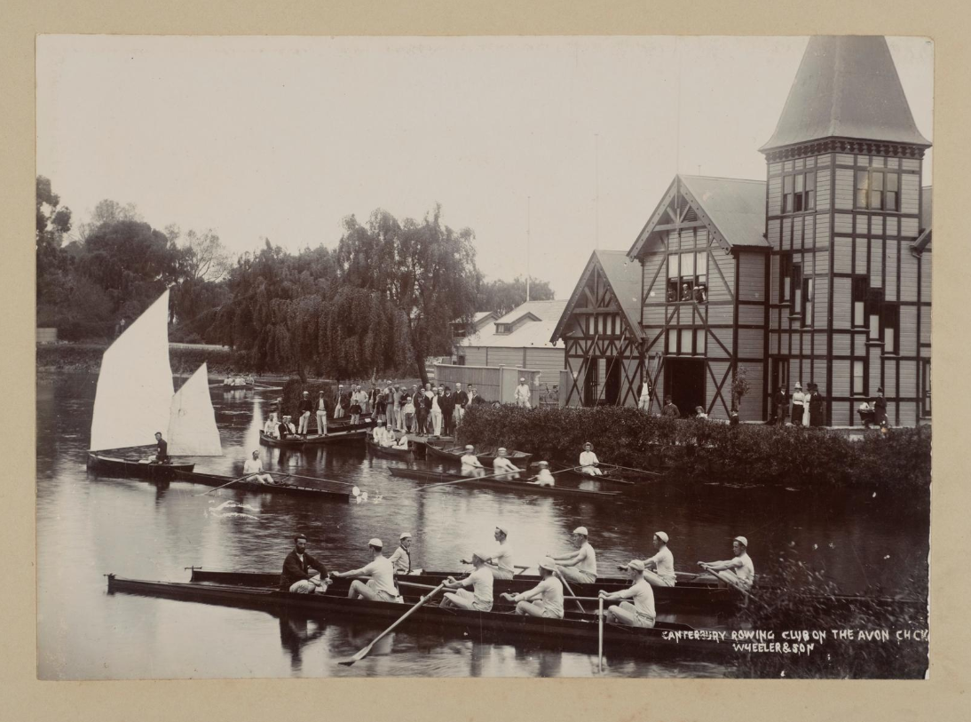 The Canterbury Rowing Club clubhouse is at left. Eight rowboats with people in them on the river (varying crew size), one empty one, one yacht with two sales. A number of people standing on the shore. Most people in the image are men.