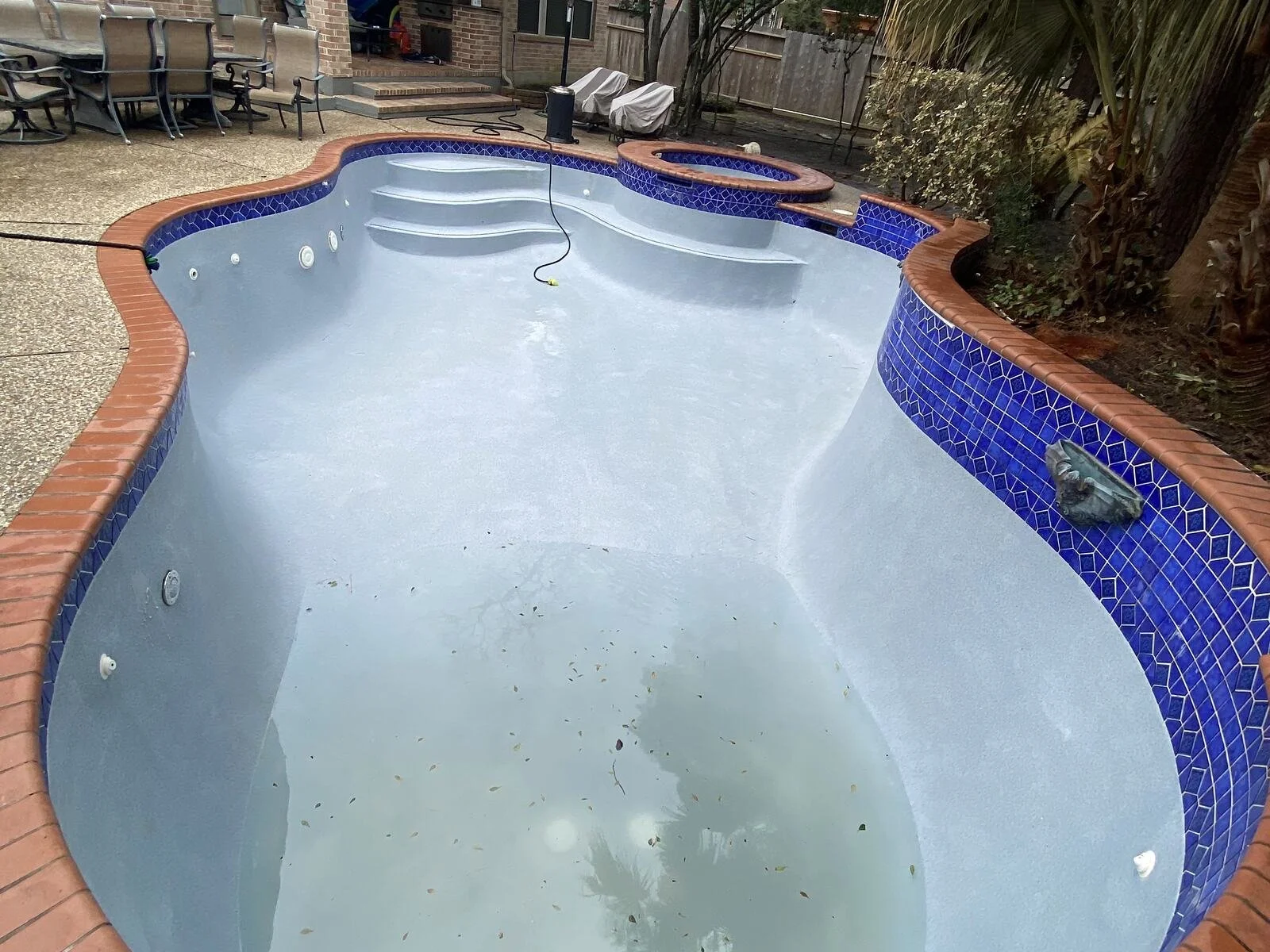 Empty in-ground swimming pool with steps and blue-tiled border, surrounded by patio chairs, covered objects, and fencing in backyard.
