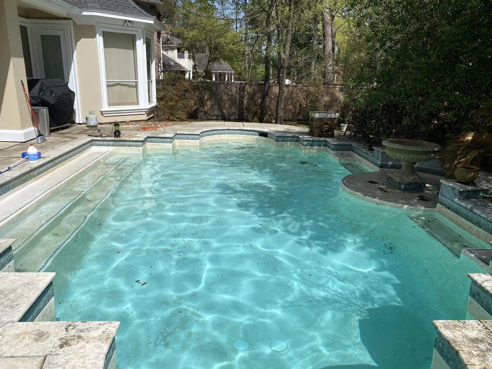 A backyard swimming pool with steps leading into the water, surrounded by a paved patio area and various outdoor items, with trees and a wooden fence in the background.