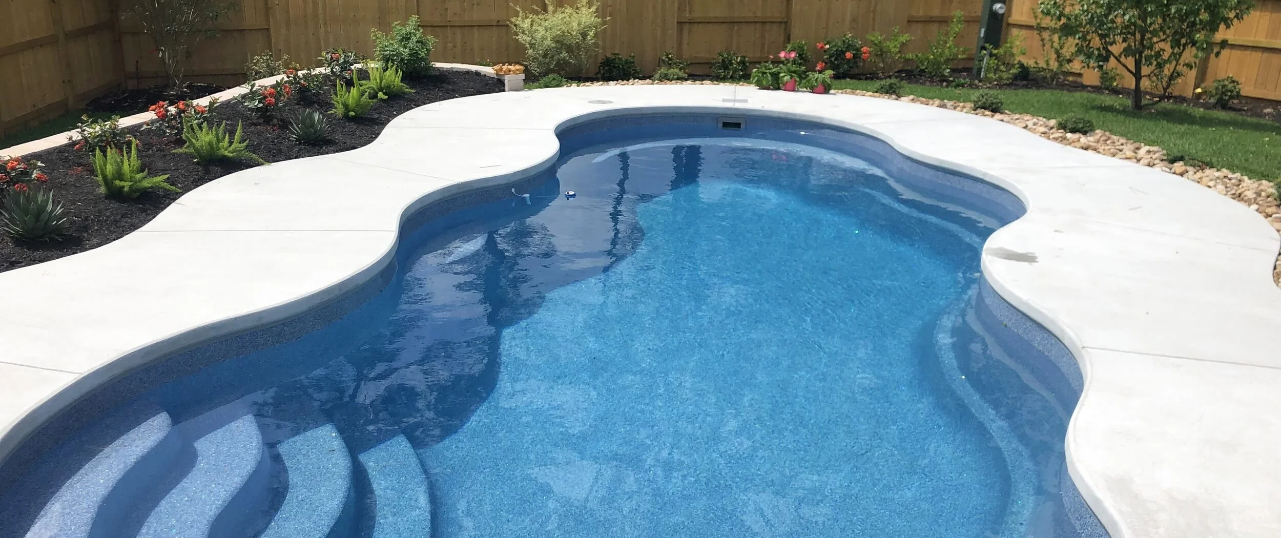 A backyard with a kidney-shaped swimming pool surrounded by a white concrete deck, with a garden bed containing plants and flowers along a wooden fence in the background.