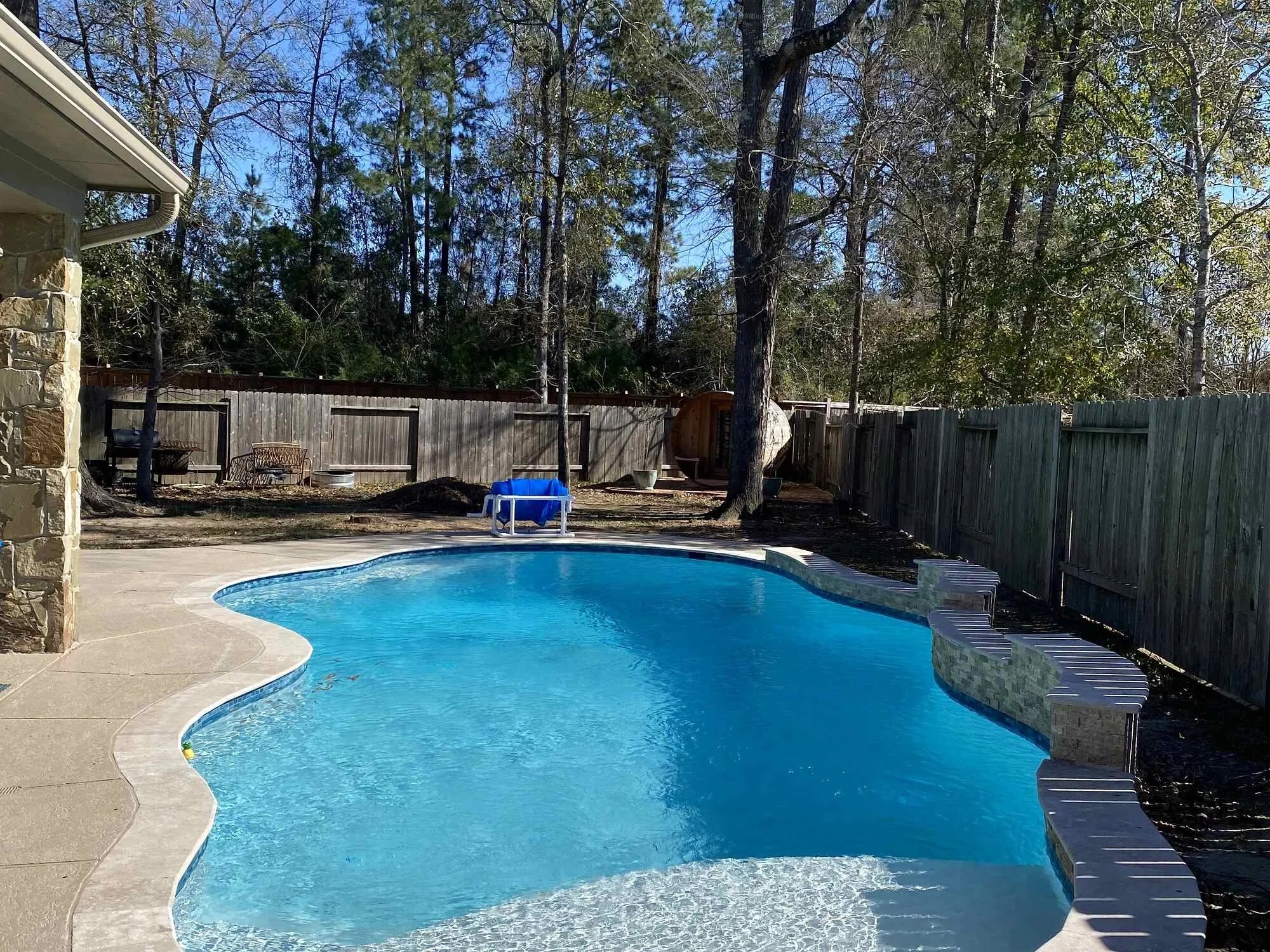 Backyard with swimming pool, surrounded by a wooden fence, trees, and a clear blue sky.