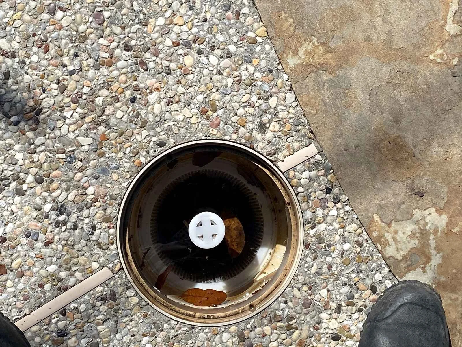 A drain cover on a concrete and pebble-textured surface, with some leaves inside the drain and a person's shoes visible at the bottom of the image.