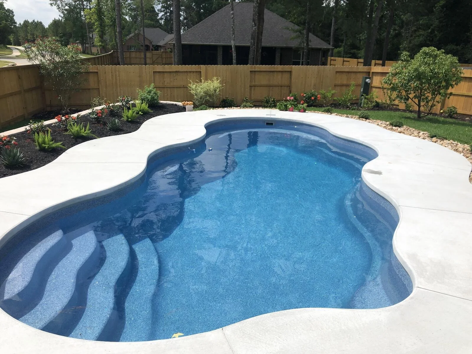 A backyard swimming pool with curved steps leading into the water, surrounded by a white concrete patio. There is a landscaped garden with plants and small bushes along a wooden fence in the background.