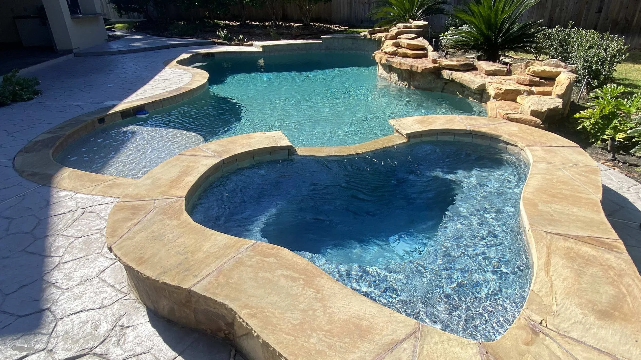 Backyard swimming pool with a built-in hot tub, surrounded by a stone deck and landscaped greenery.