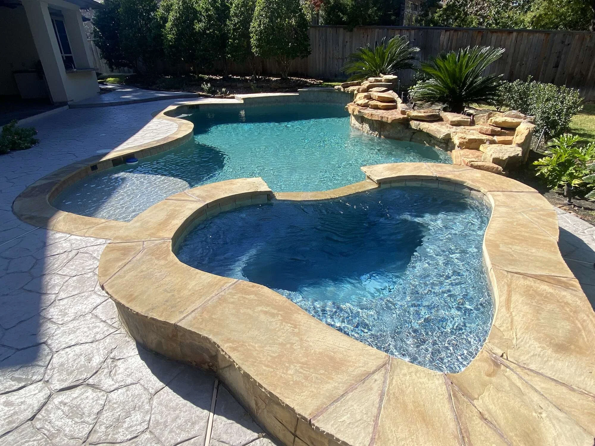 A backyard swimming pool with a connected hot tub, surrounded by stone paving and landscaped with bushes and large plants, enclosed by a wooden fence.
