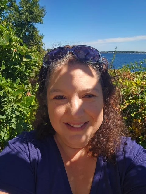 A woman with curly hair wearing sunglasses takes a selfie outdoors, with greenery and a body of water in the background on a sunny day.