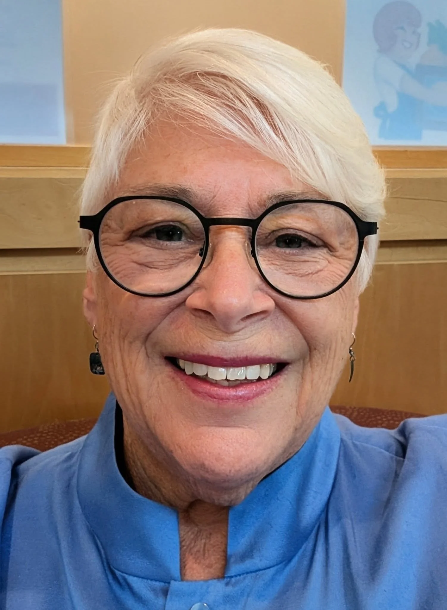 Close-up of an elderly woman with short white hair, wearing glasses and a blue top, smiling in a room with light-colored walls and a wooden background.