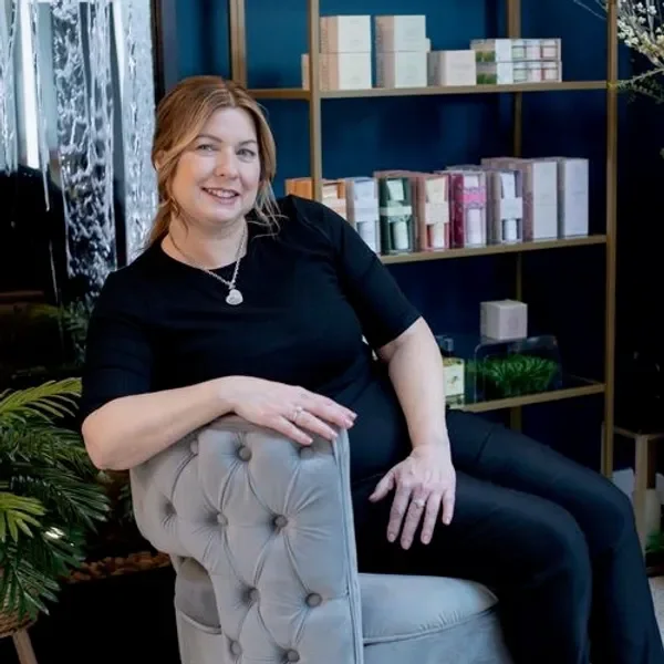 A woman with shoulder-length reddish hair, wearing a black top and necklace, sitting on a beige tufted armchair, smiling in an interior space with a bookshelf filled with pastel-colored boxes and a dark blue wall in the background.