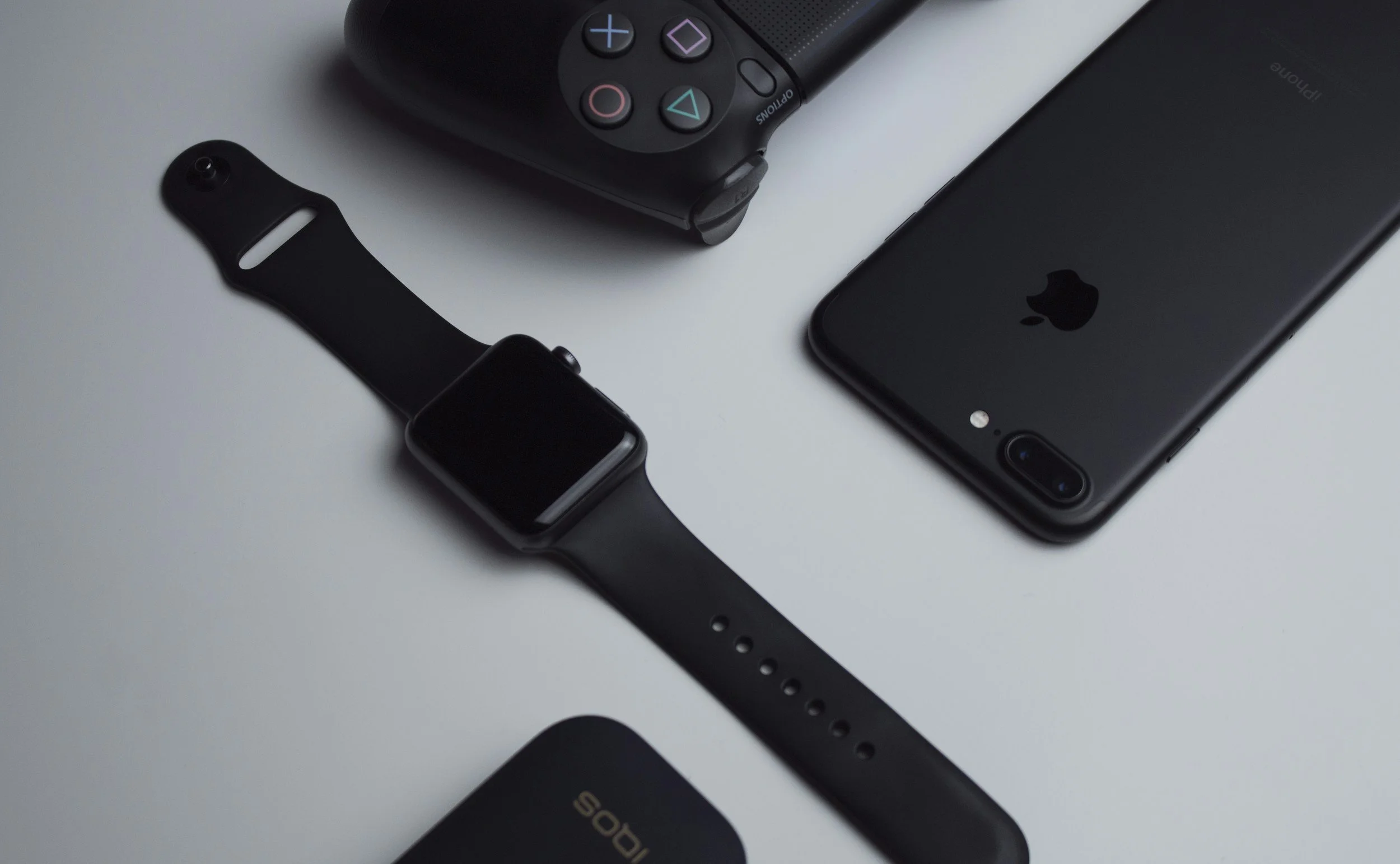 Collection of electronic devices including a black PlayStation 4 controller, a smartwatch, a black iPhone, and a black iPad placed on a light gray surface.