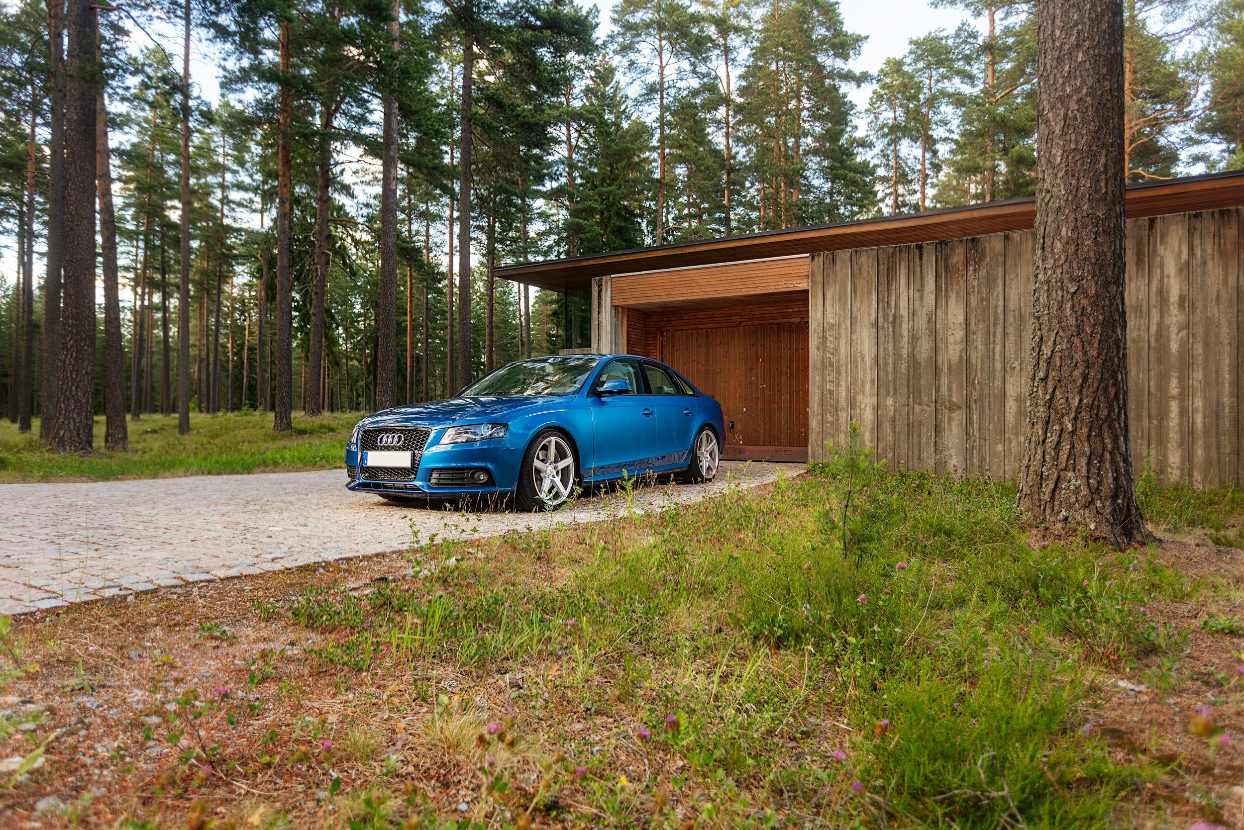 A blue Audi sedan parked on a gravel driveway in front of a modern wooden house surrounded by tall pine trees.
