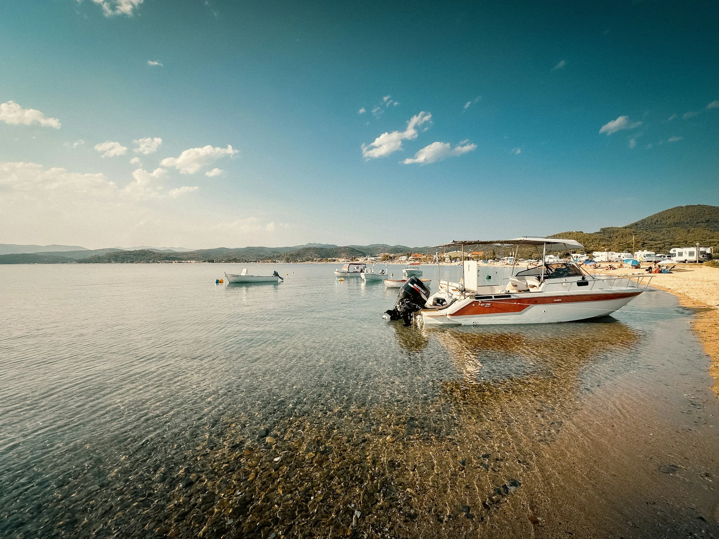 A peaceful lakeside scene with boats floating on calm water, a pebble shore, and a backdrop of hills under a partly cloudy sky.