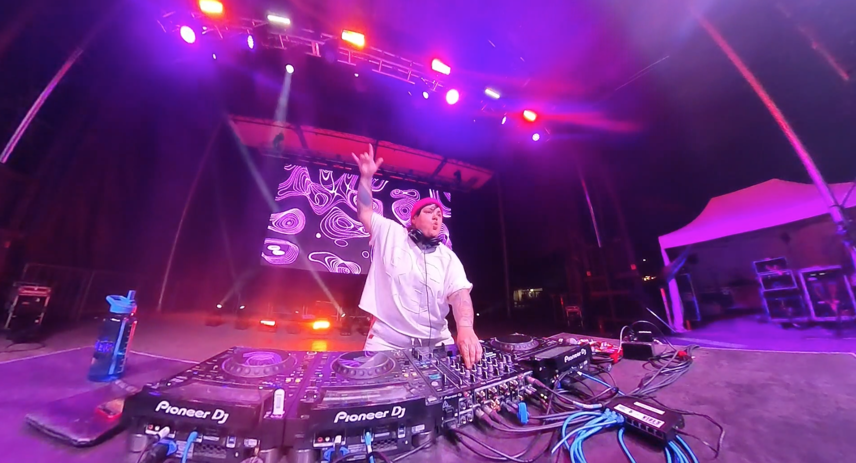 A DJ performing on stage at a concert with colorful lighting and a digital backdrop, wearing headphones and a white outfit, raising one hand.
