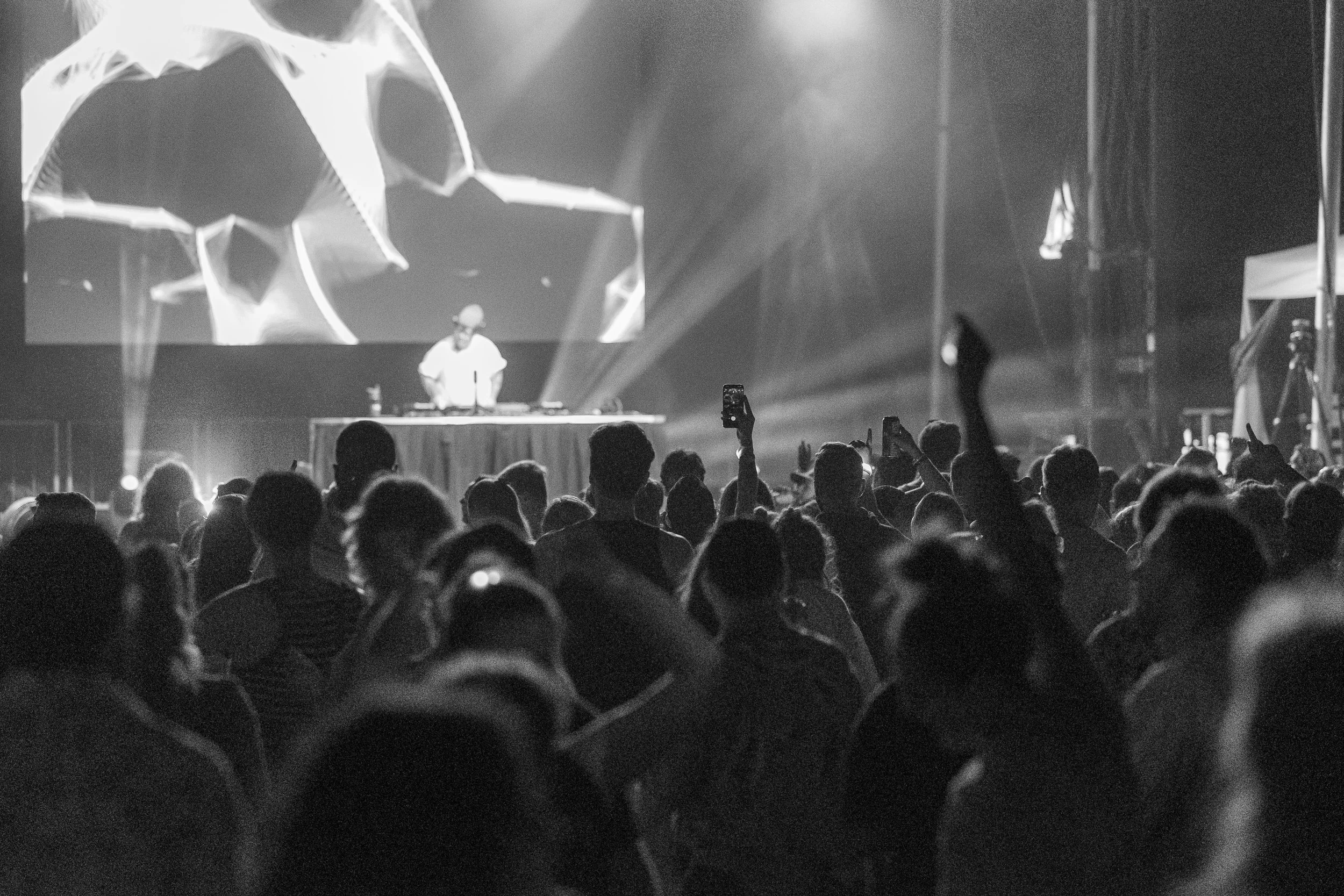 A black and white photo of a concert with a crowd of people dancing and a DJ on stage. The DJ is wearing a white shirt and standing behind turntables. There are bright stage lights and a large screen displaying abstract visuals in the background.