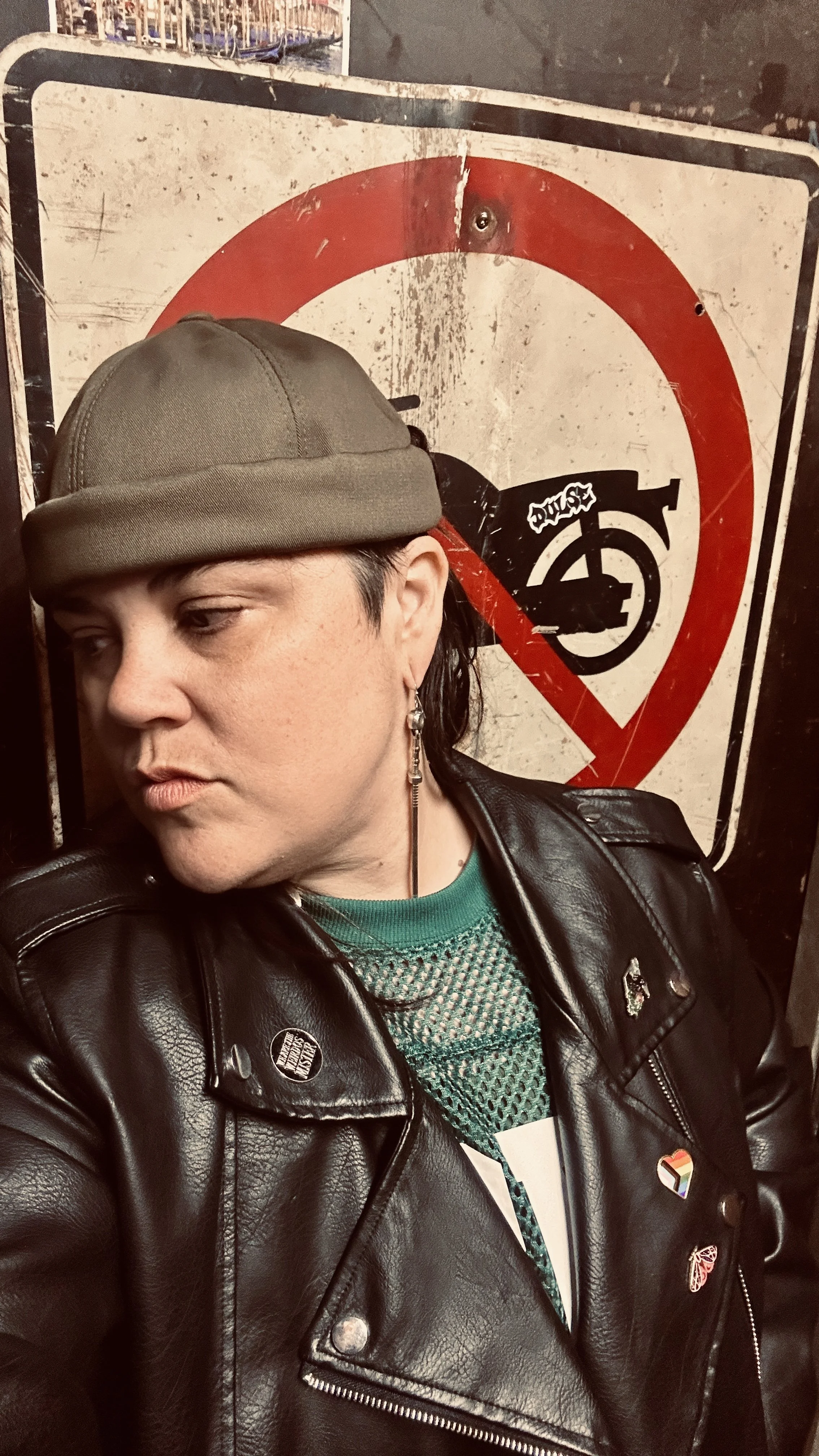 A woman taking a selfie in front of a weathered no skateboarding sign. She is wearing a leather jacket, a green top, and a hat, with various pins and rainbow-themed badges on her jacket.