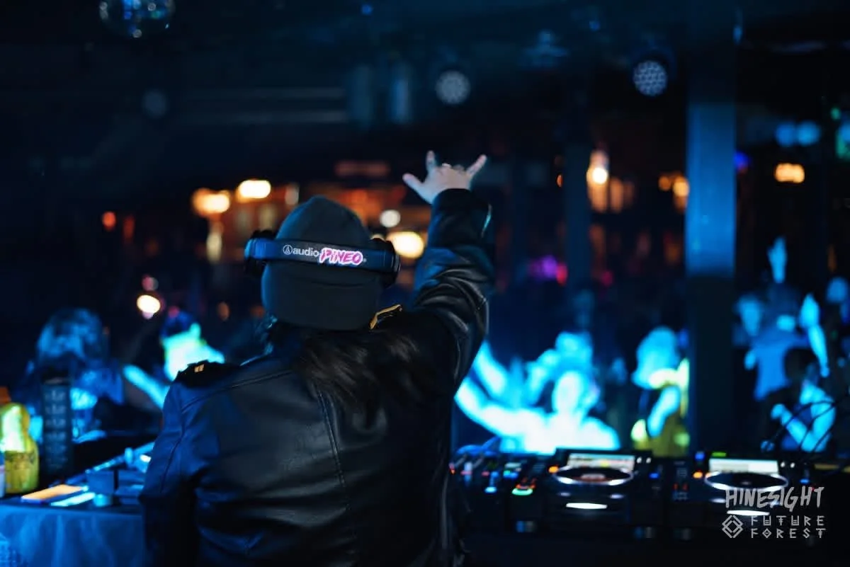 A DJ wearing a black beanie and headphones with the logo PINEO, performing at a nightclub or concert with a crowded, illuminated dance floor in the background.