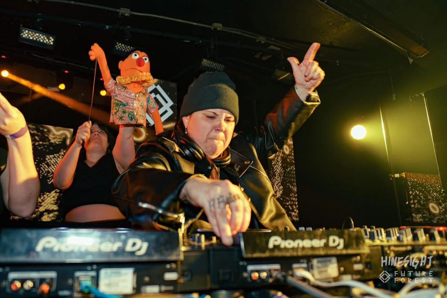 DJ in a beanie and leather jacket playing music at a club with a puppet character in a Hawaiian shirt and leis, raised in the background with party atmosphere lighting.