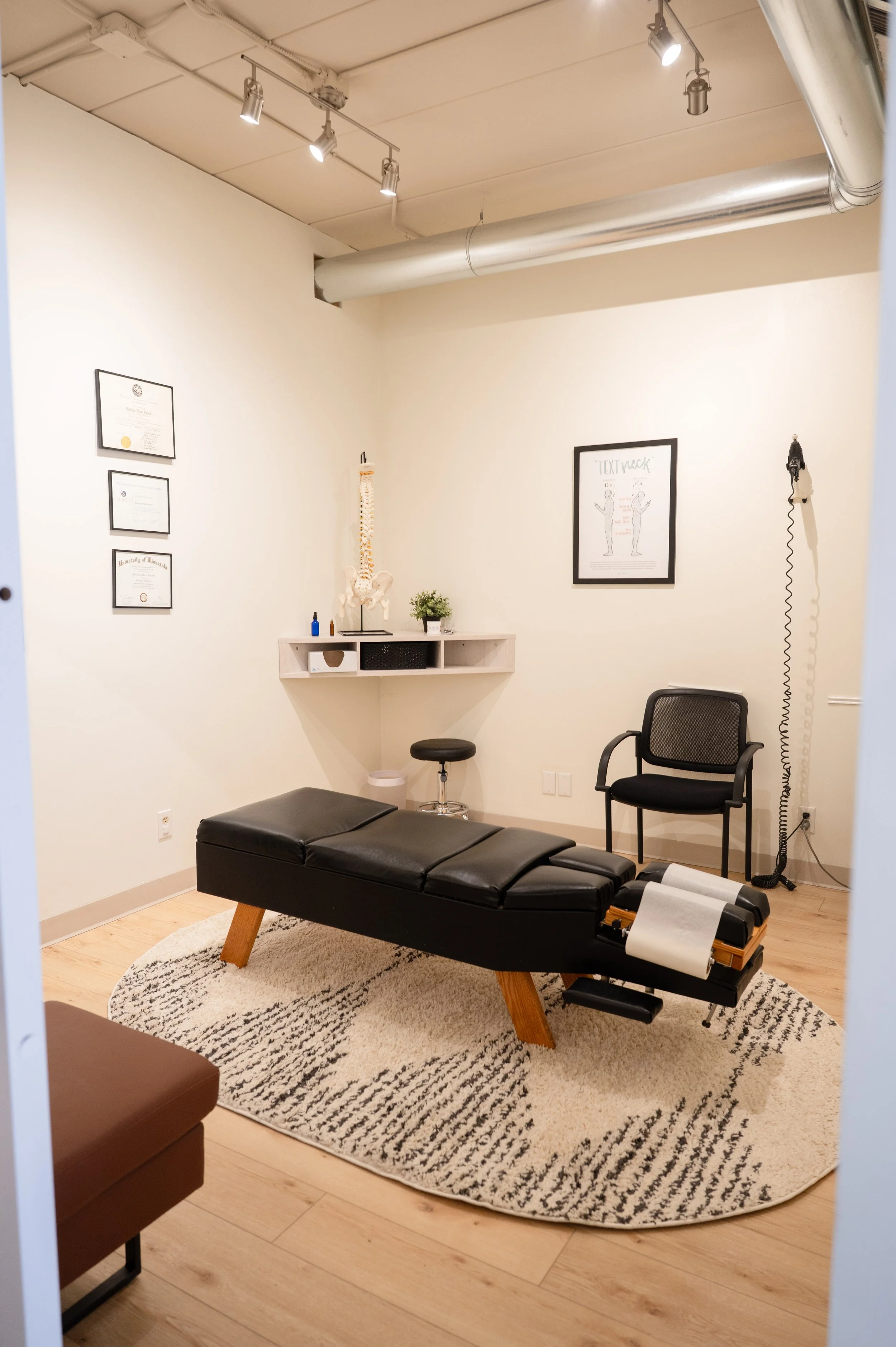 A medical or chiropractic office treatment room with a black padded examination table and a black chair. The room has a cream-colored rug, a wall-mounted shelf with a spine model, framed diagrams, and certificates. There is a wall-mounted electrical device with a coiled cord on the right.