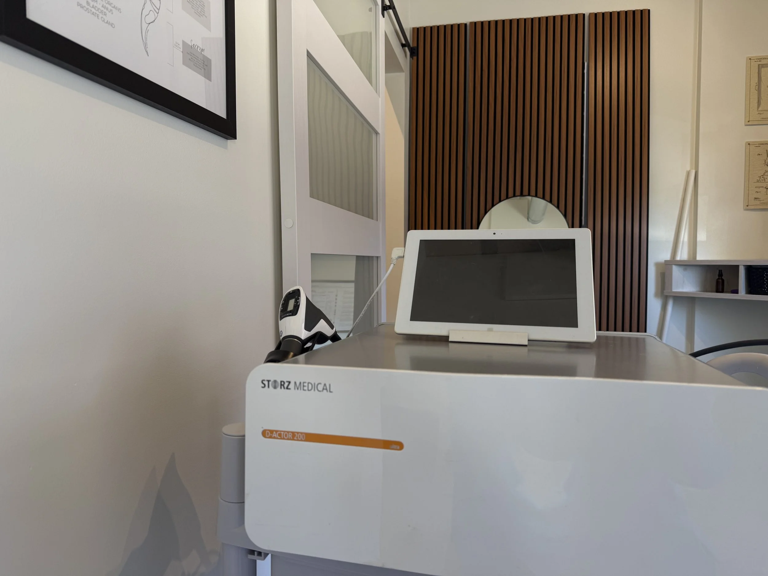 Medical office or clinic workspace with a white desk, medical device labeled 'D-ACTOR 200', and a tablet or computer monitor. The background has a wooden slat wall and white railing. There are framed diagrams on the walls.