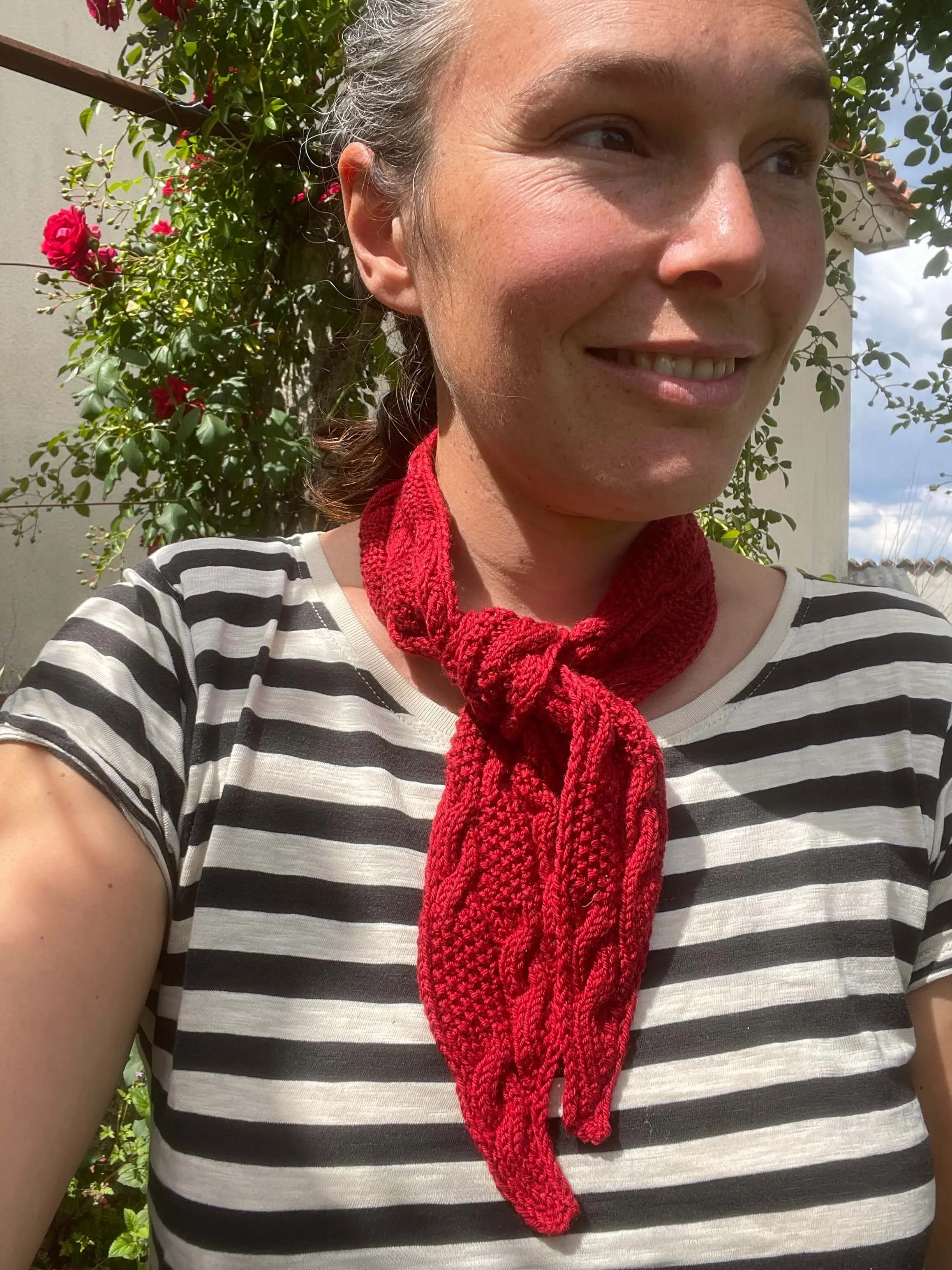 A woman wearing a red knitted scarf is smiling next to some flowers. The mini scarf has cables and a moss stitch design.