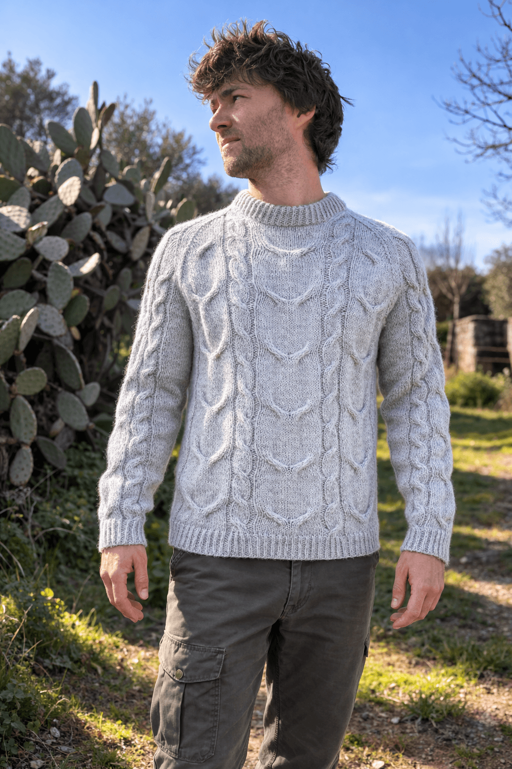 A man stands outdoors during daytime, wearing a light gray knitted sweater with a cable pattern. There are cactus plants in the background and blue sky. The jumper is handmade using a modern knitting pattern for men available for instant pdf download