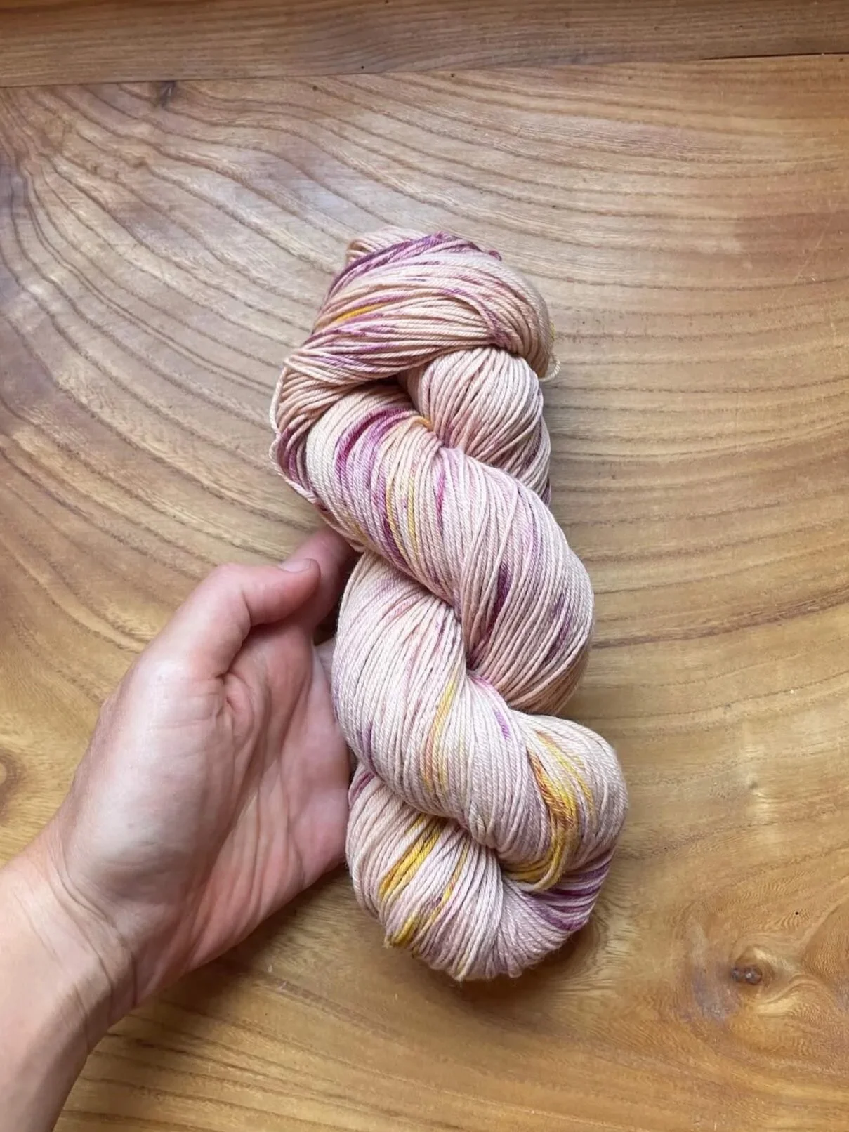 A skein of light pink yarn with yellow and darker pink specks, held in a person’s hand, resting on a wooden surface.