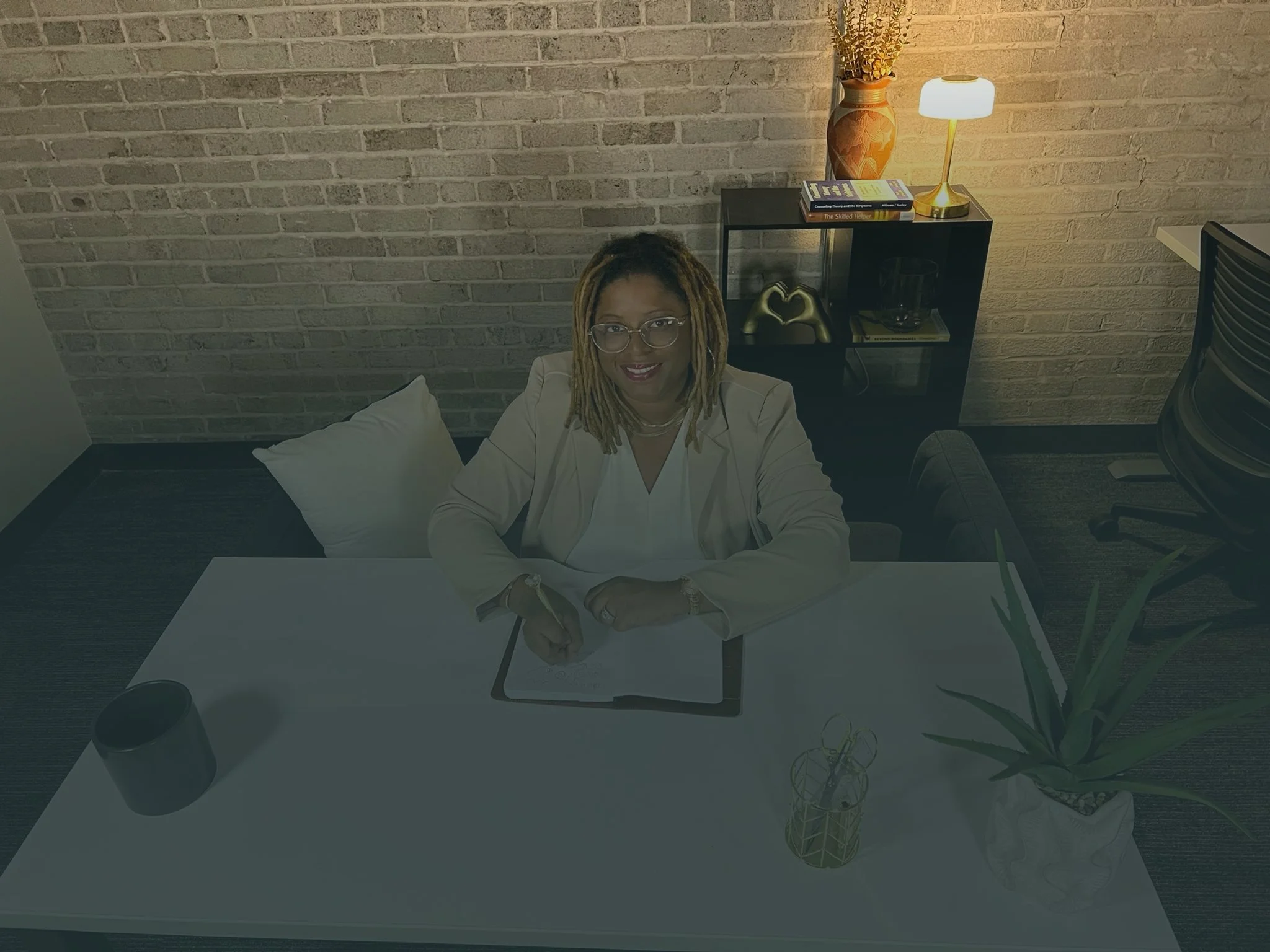 A woman with dreadlocks and glasses sitting at a white desk in a modern office space, smiling at the camera. Behind her is a brick wall, a black bookshelf with books and decor items, and a small gold and white table lamp with a floral vase on top. There are also a plant, an open laptop, a coffee cup, and some stationery on the desk.