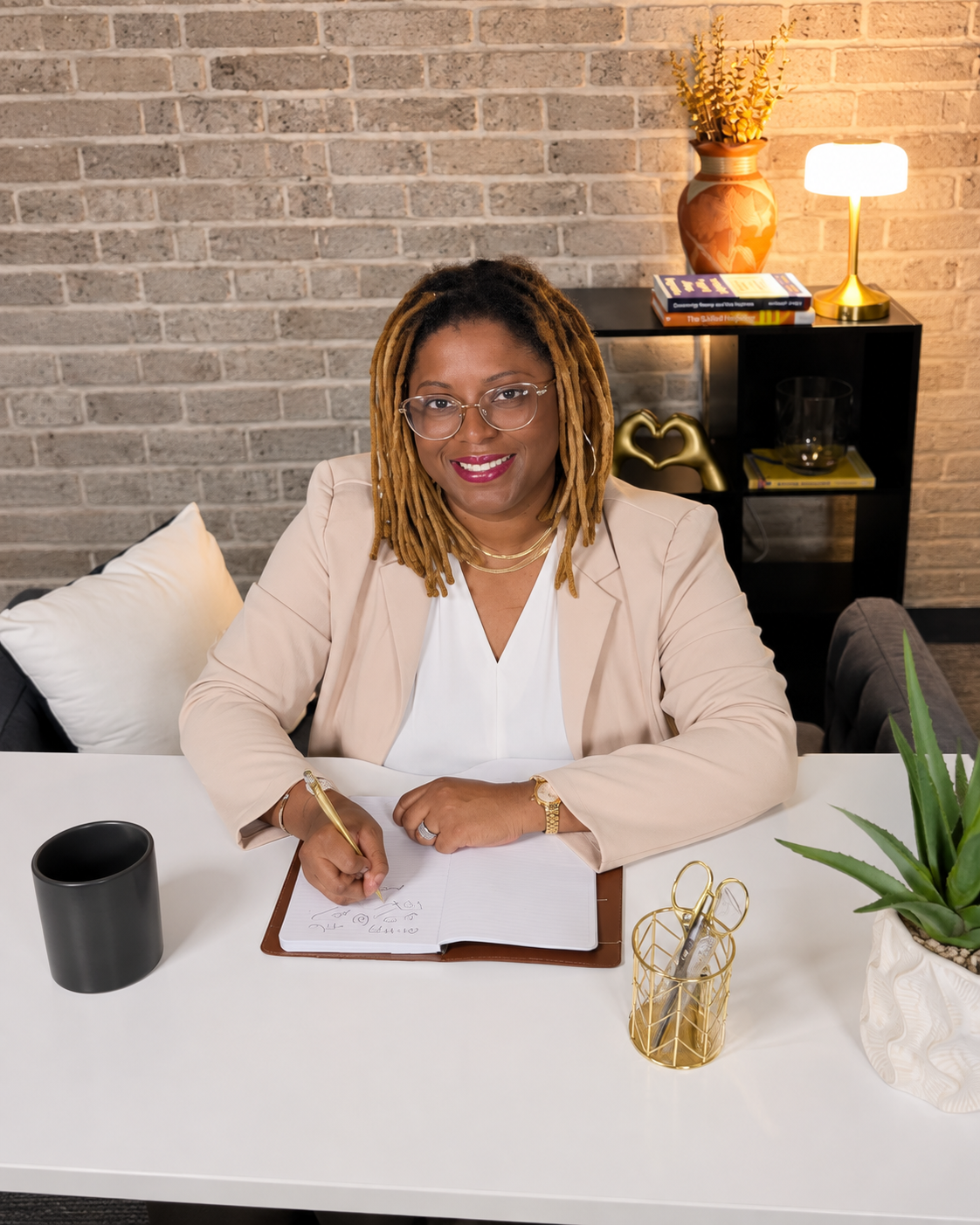A woman with glasses and dreadlocks sitting at a white desk, smiling, with a black mug and a gold pen holder containing scissors on the desk, a notebook in front of her, and a potted plant to the right; a brick wall and a shelf with books, a flower vase, a gold heart sculpture, and a table lamp in the background.