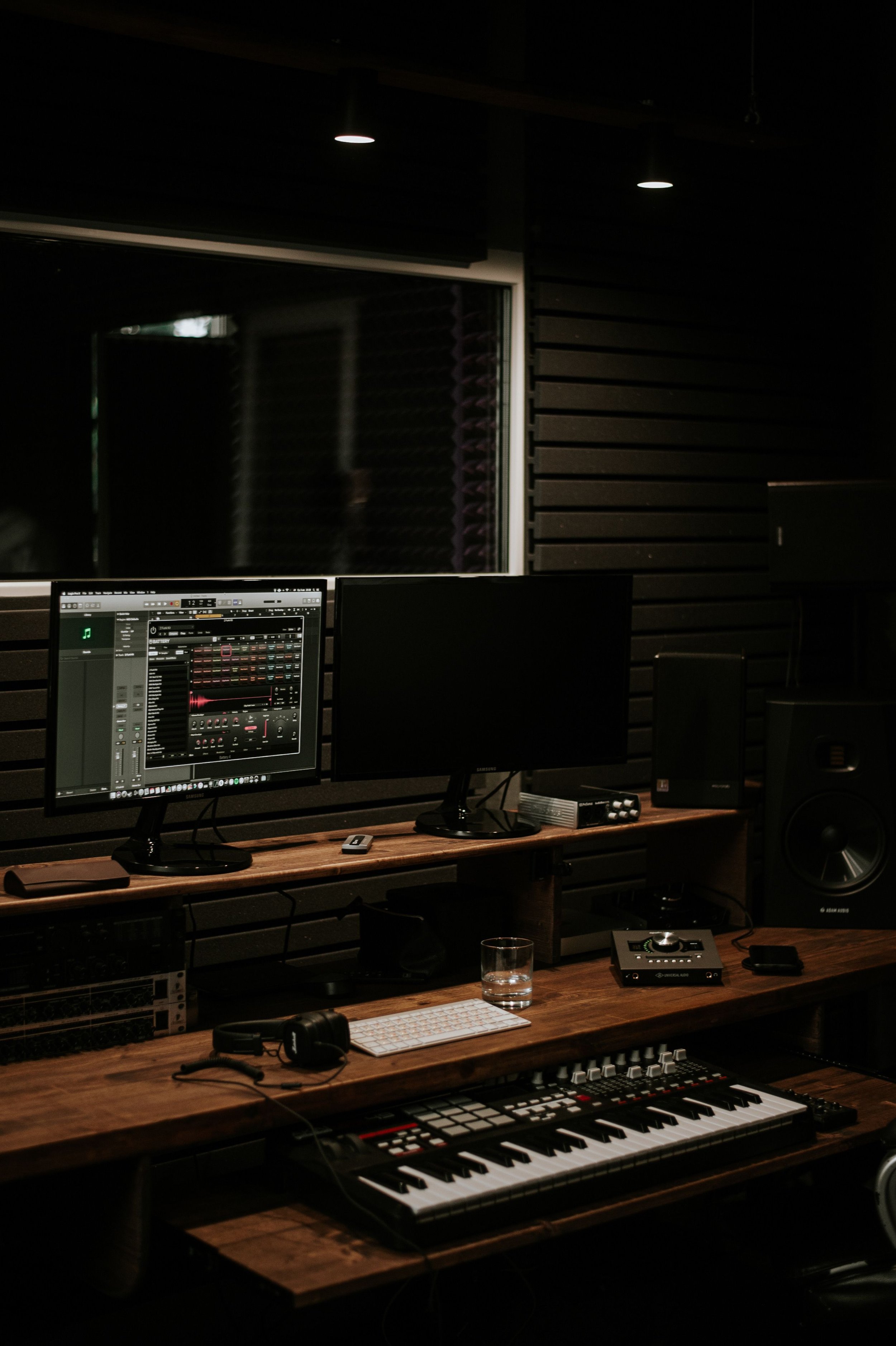 Recording studio with multiple computer monitors, sound equipment, a keyboard, and a glass of water on a wooden desk, surrounded by acoustic foam panels on the walls.