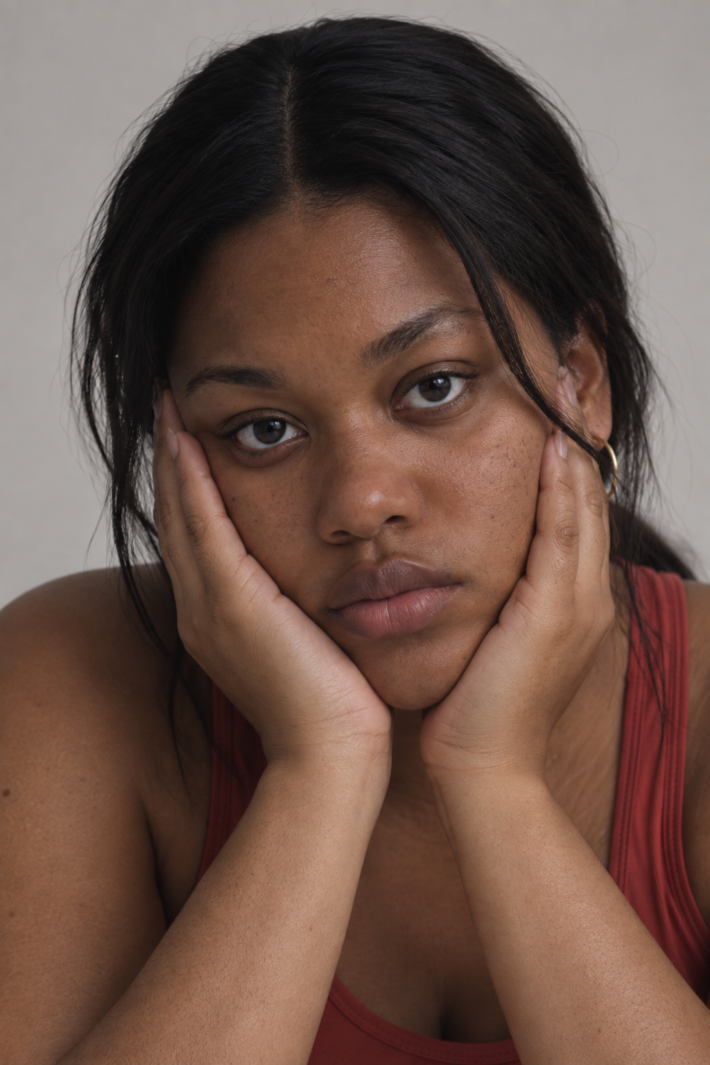 Close-up of a woman with dark hair, resting her face in her hands, wearing a red tank top.
