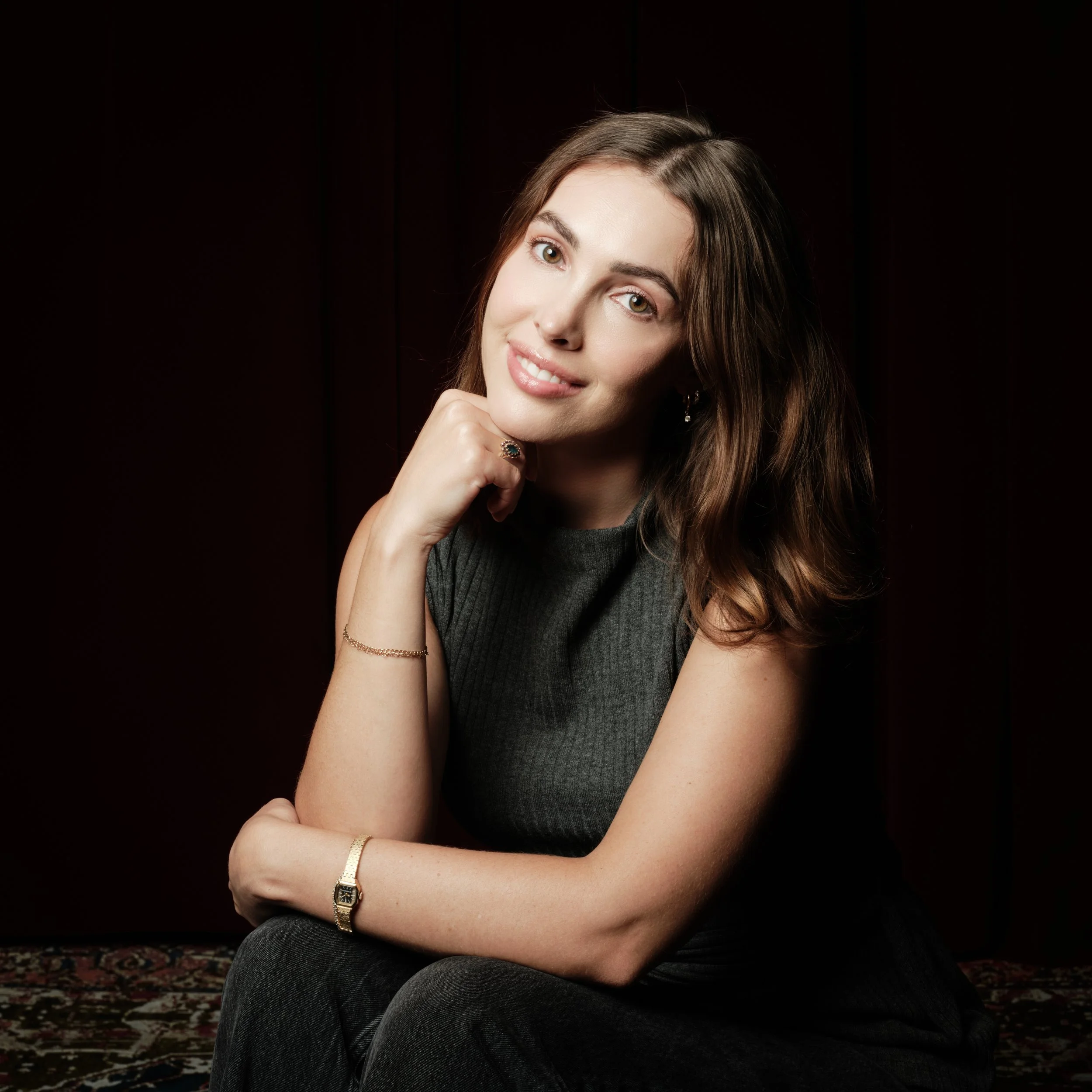 Portrait of a young woman with brown hair, green eyes, smiling slightly, wearing jewelry, sitting with her arms crossed against a dark background.