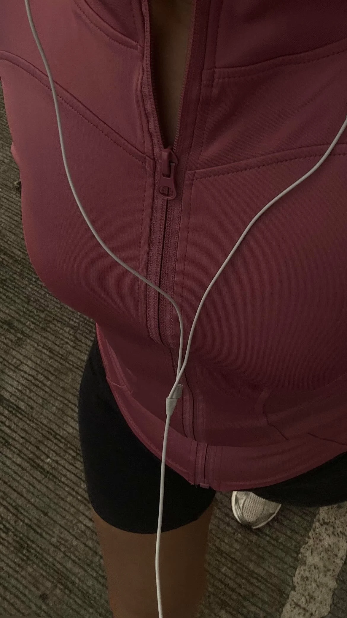 Close-up of a person wearing a pink jacket with a zipper, black shorts, and white sneakers, standing on a carpeted floor, with white earphones hanging down.