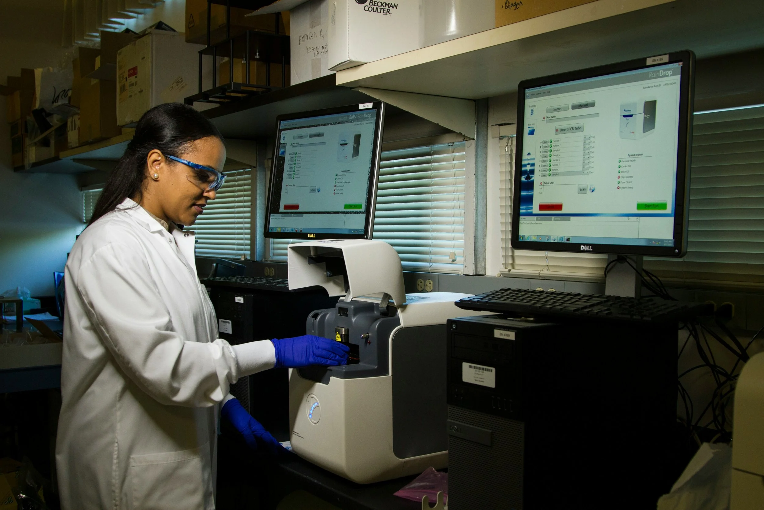 A scientist in a white lab coat, safety glasses, and blue gloves working with a laboratory instrument in a research lab with computer monitors displaying laboratory data.