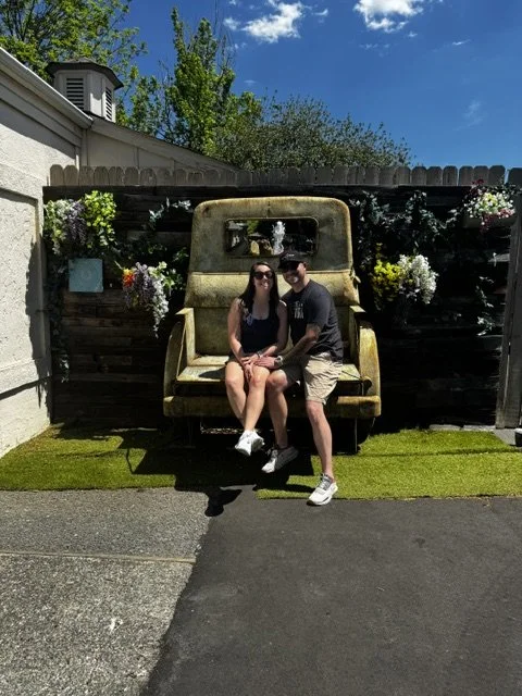 Two people sitting on a vintage yellow car seat bench in a backyard with flowers on a wooden fence and a clear blue sky.