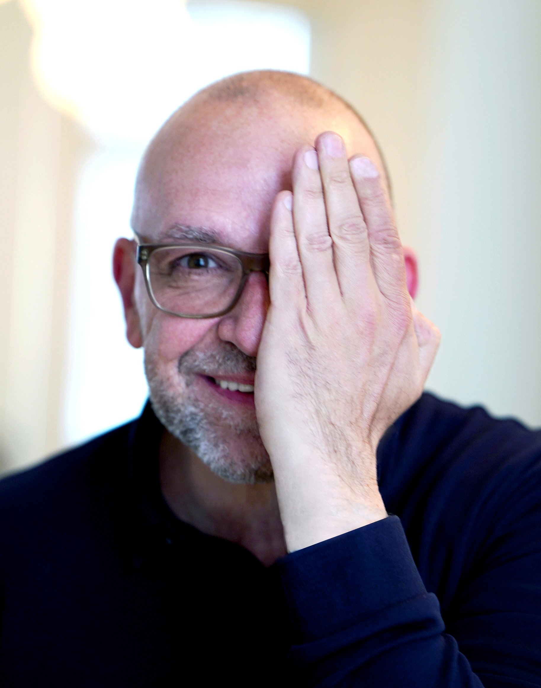 Ein Mann mit Glatze, Brille und grauem Bart hält eine Hand vor sein Gesicht, bedeckt die rechte Augenhälfte, leicht lächelnd, im Hintergrund unscharfer heller Raum.