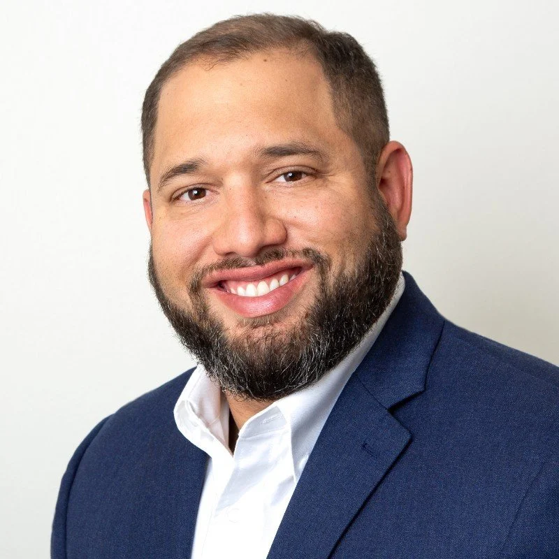 Headshot of a man with short brown hair and a beard, smiling, wearing a navy blue suit jacket and white shirt against a plain white background.