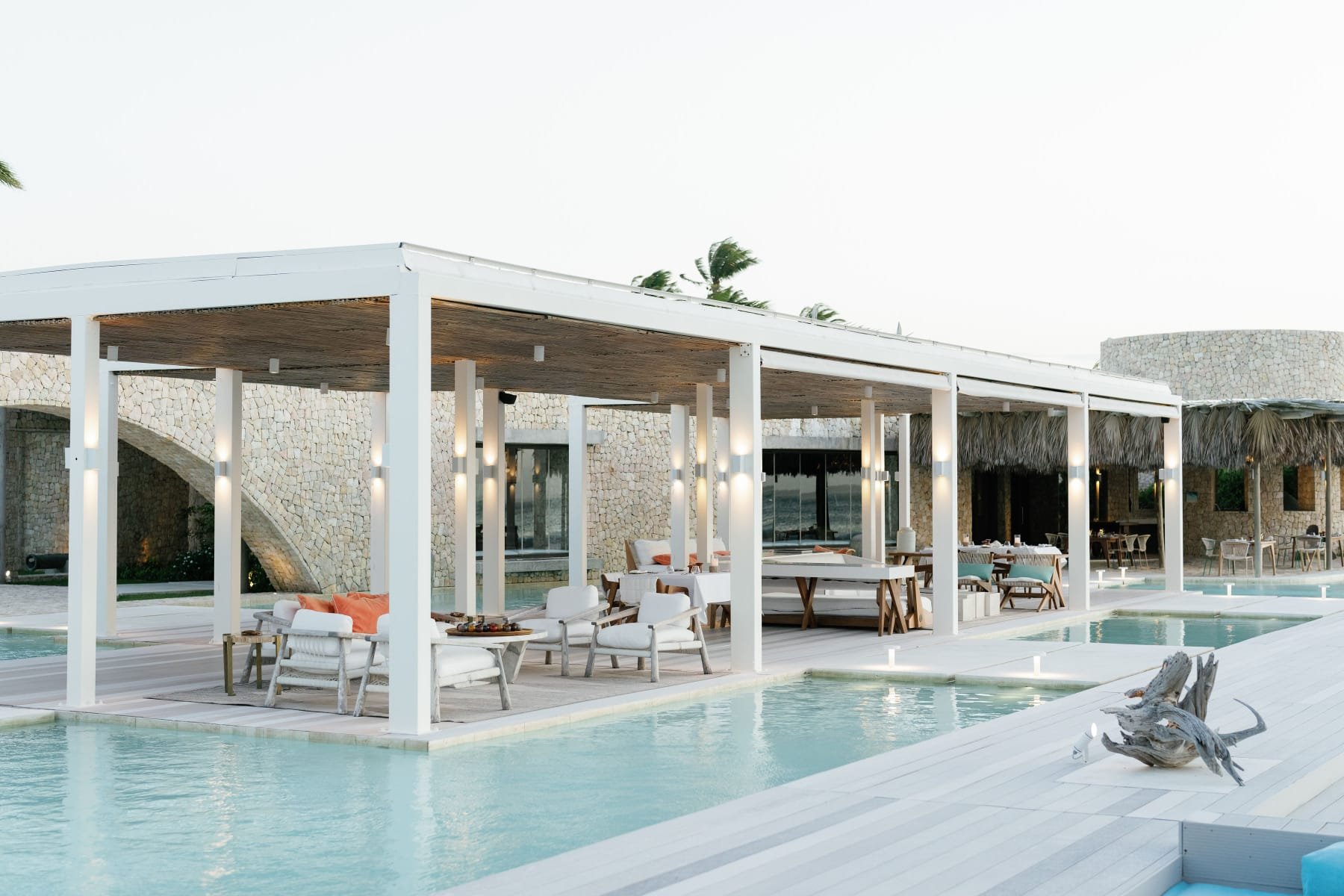 Ein moderner, luxuriöser Poolbereich im Freien mit weißen Sonnenschirmen und mediterraner Architektur. Es gibt Sitzgelegenheiten, Tische und Liegen neben einem Pool, mit Sanddünen und Wind im Hintergrund.