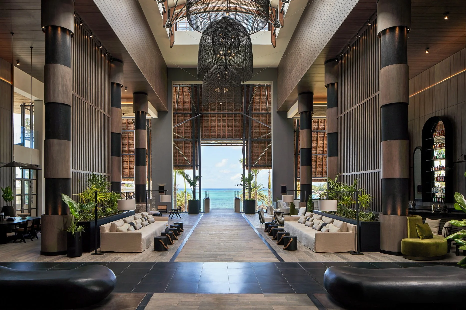 Innenraum eines modernen Resorts Hotels mit Blick auf den Strand und das Meer im Hintergrund, große schwarze Lüster, Holz- und Pflanzenakzente, helle Sitzgelegenheiten und eine offene, elegante Atmosphäre.