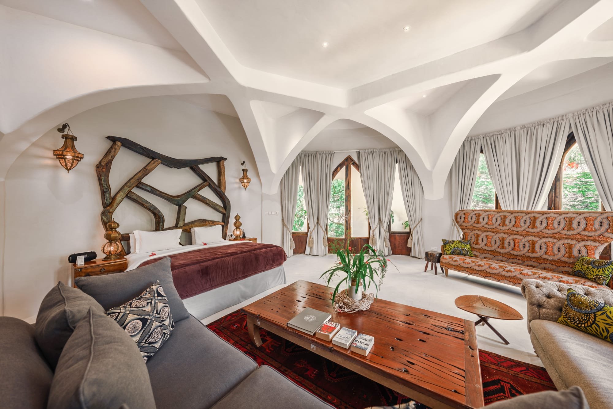 Ein geräumiges Wohnzimmer mit hohen weißen Decken, großen Fenstern mit weißen Vorhängen, einem großen grauen Sofa mit Kissen, einem rustikalen Holztisch mit Büchern, einem langen braunen Sofa mit türkisen Kissen und einem großen, gepolsterten Stuhl mit buntem Muster.
