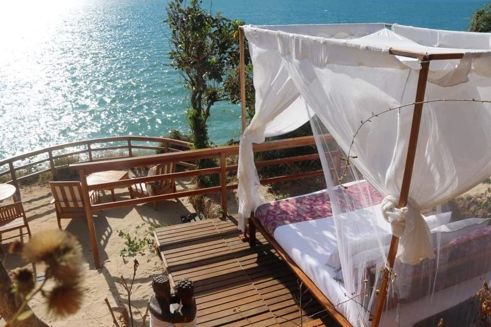 Ein Apparat, der auf einem Balkon am Meer steht. Das Bett hat ein weißes Tuch und sieht einladend aus. Es ist sonnig und es gibt ein paar Stühle und Tische auf dem Balkon.