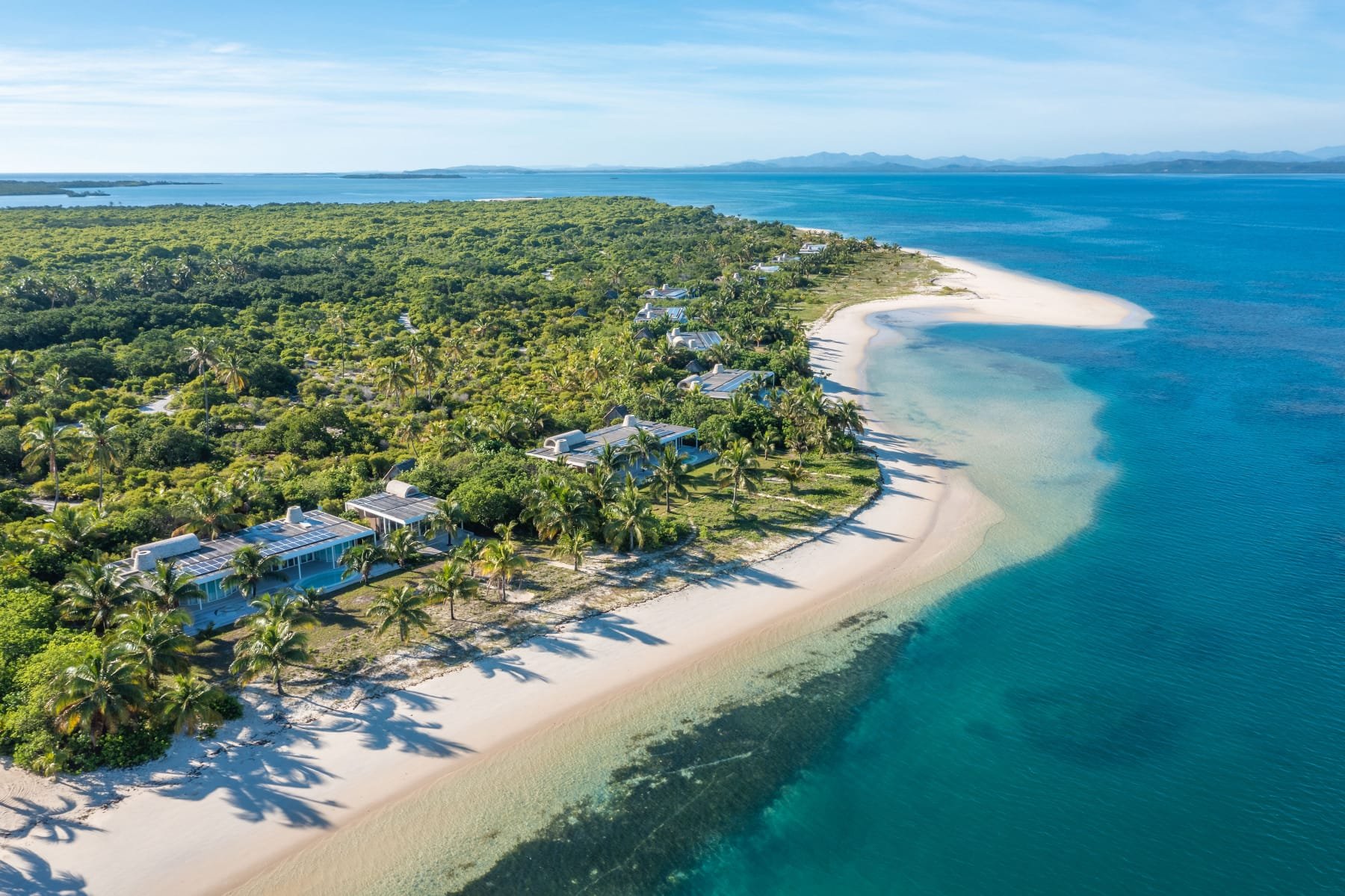 Luftaufnahme eines Strandes mit weißen Sand, üppigem Grün und modernen Resorts am Meer, mit klarem Wasser und Hügel im Hintergrund.