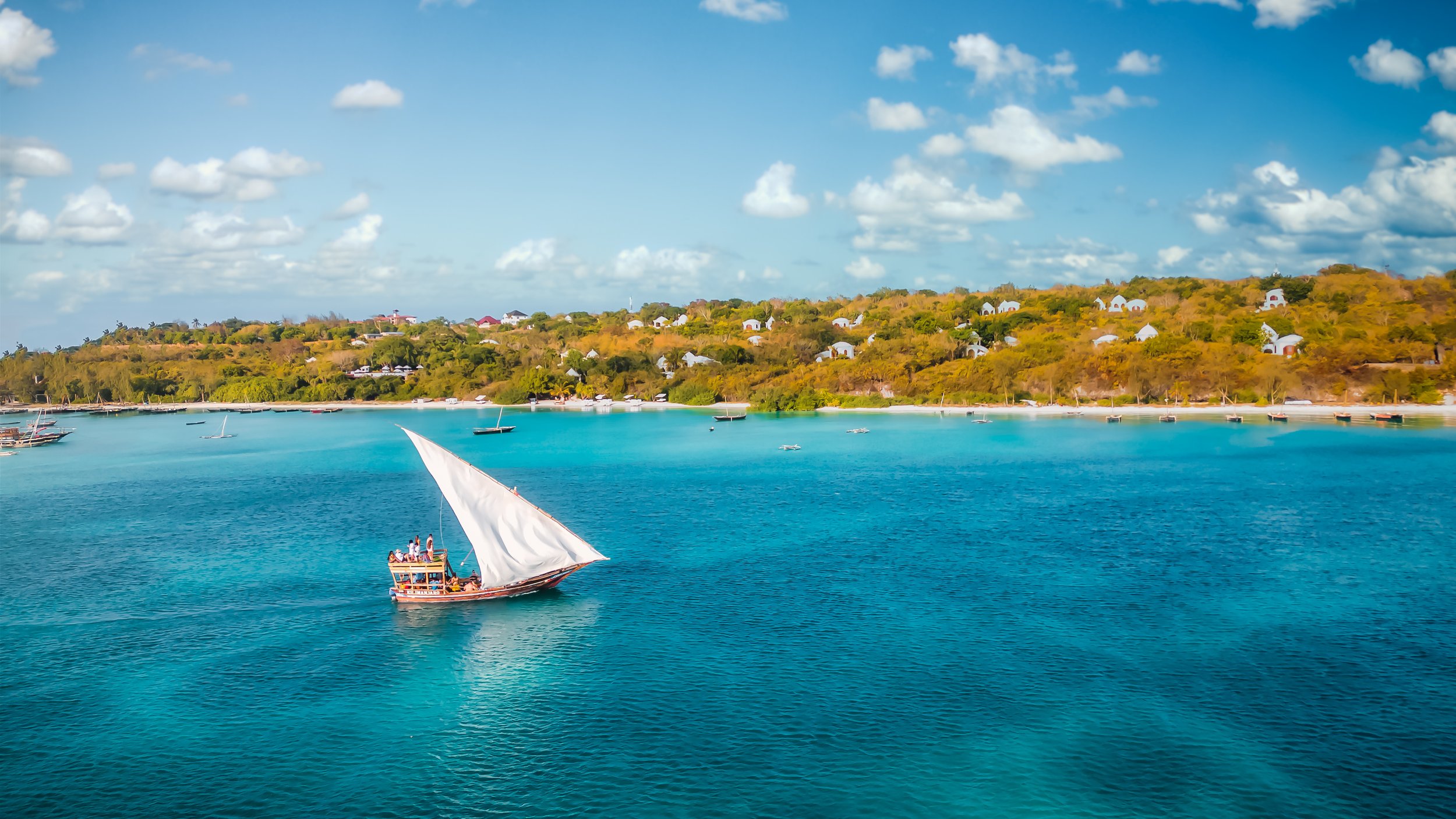 Blick auf eine türkisblaue Bucht mit einem traditionellen Segelboot und mehreren kleineren Booten, im Hintergrund eine bewaldete Hügelkette mit weißen Gebäuden unter einem blauen Himmel mit Wolken.