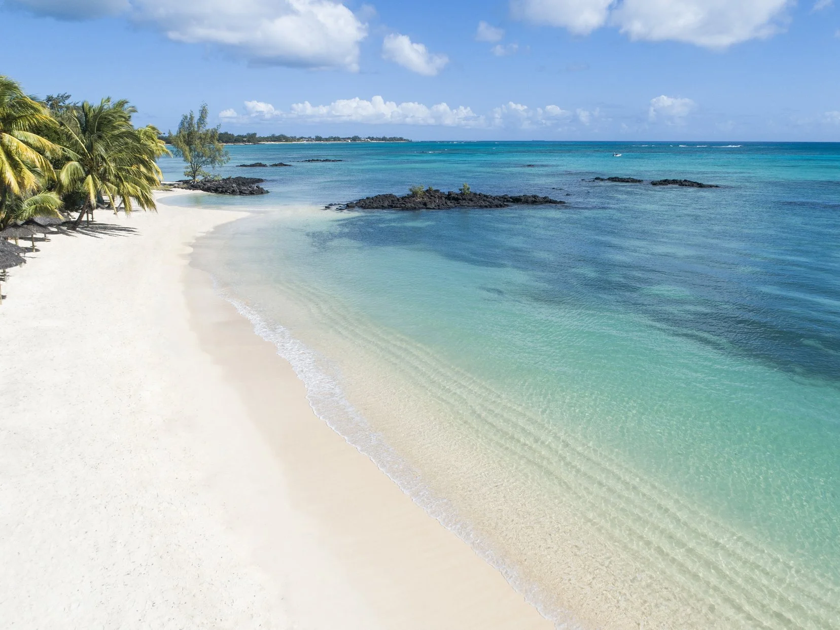 Strand mit weißen Sand und Palmen, ruhiges türkisfarbenes Meer unter blauem Himmel mit Wolken, fernen Land im Hintergrund.