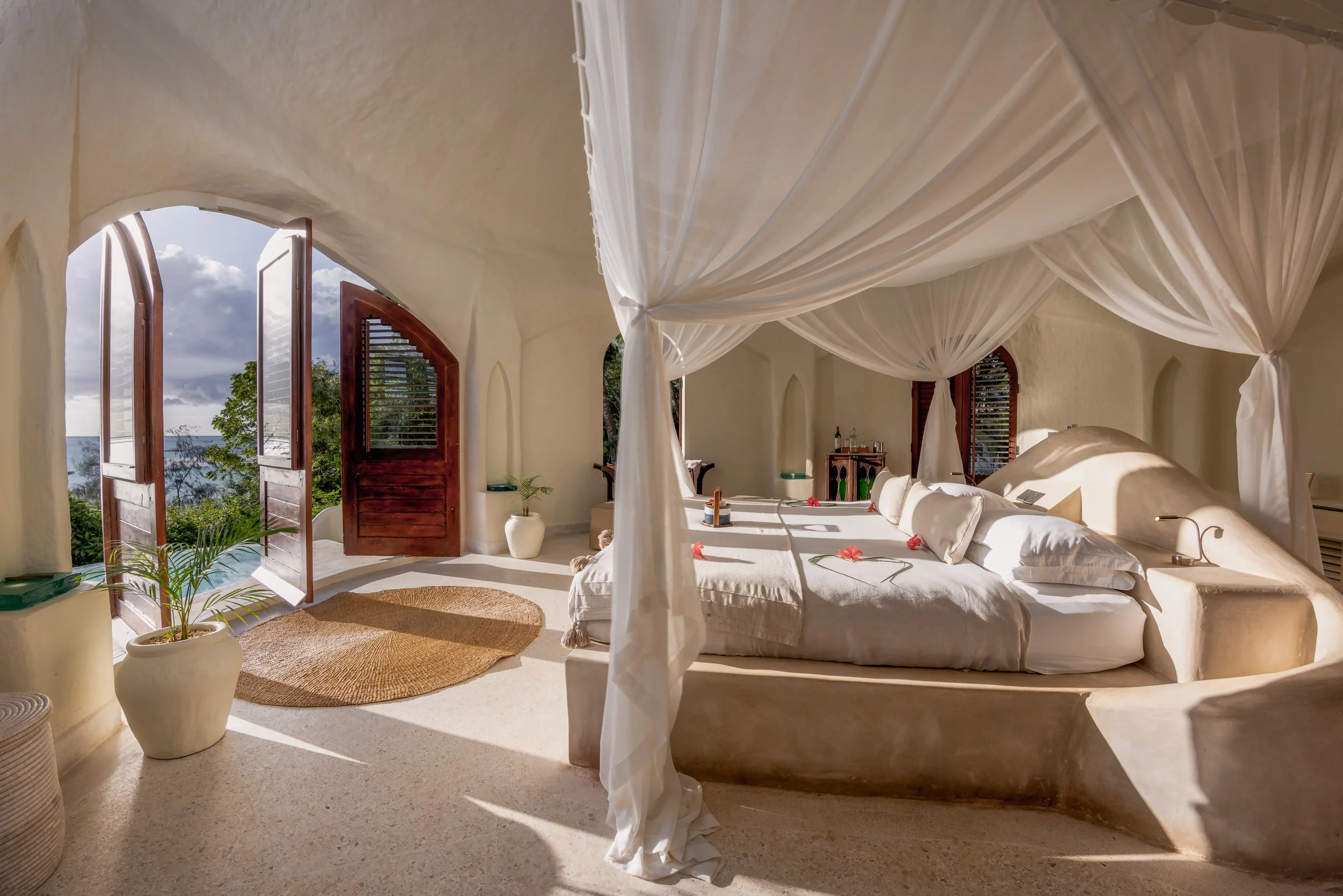 Ein Schlafzimmer mit weißen Vorhängen, großen offenen Fenstern mit Blick aufs Meer, Schlafzimmermöbeln und Dekoration im entspannenden Stil.