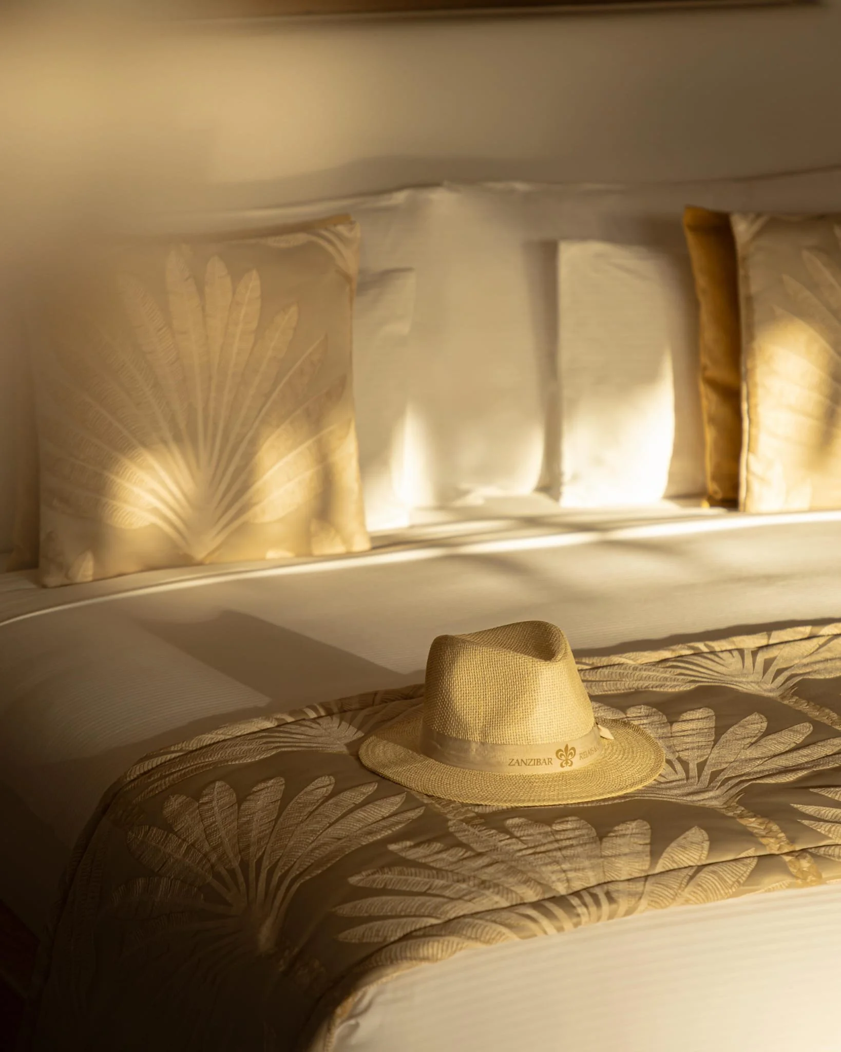 Ein Bett mit Bettwäsche, zwei Kissen mit goldfarbenen Kissenbezügen, und ein Strohhut mit der Aufschrift 'ZANZIBAR'.
